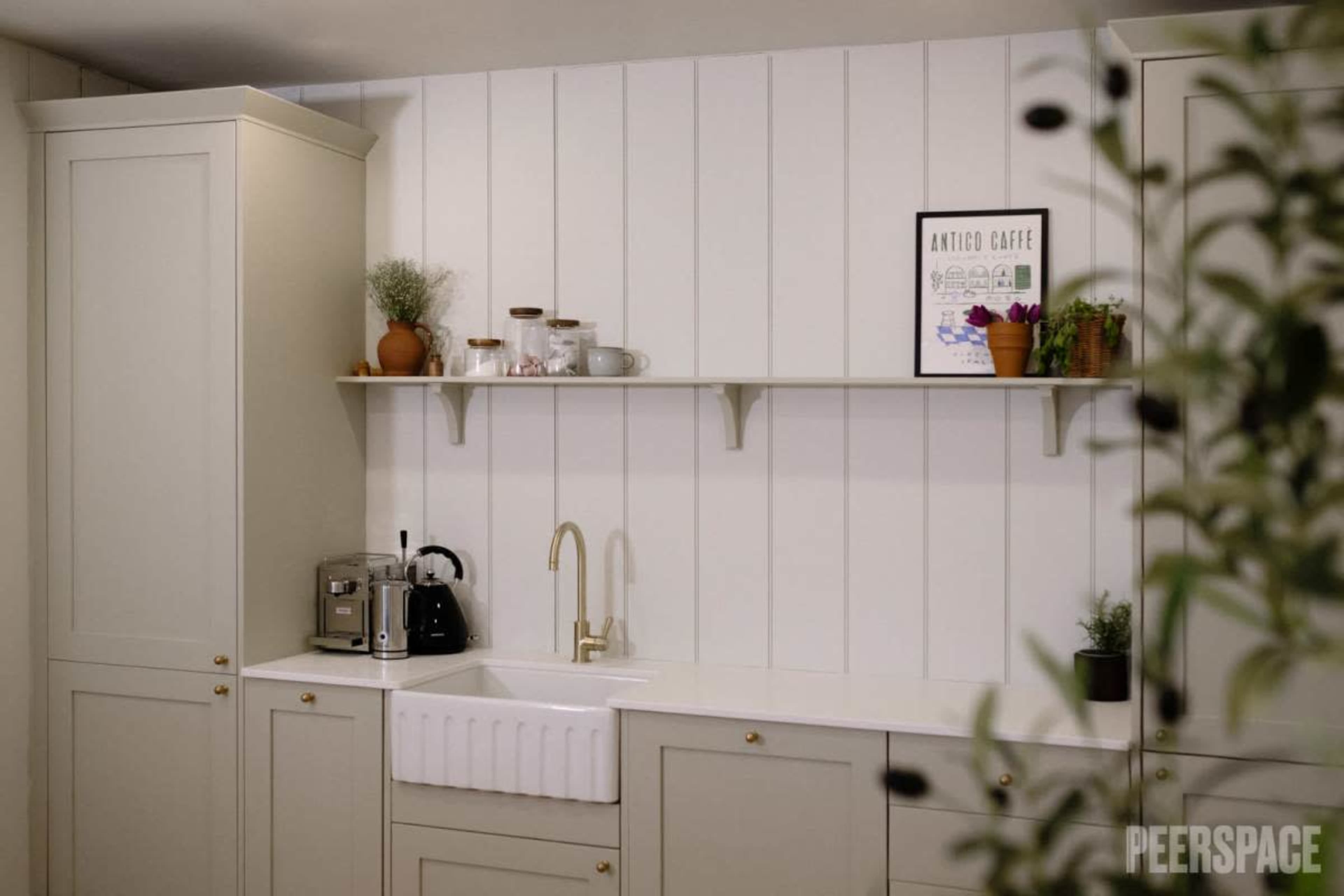 A modern kitchen with light-colored cabinetry, a farmhouse sink, and a shelf displaying plants and kitchenware against a paneled wall.