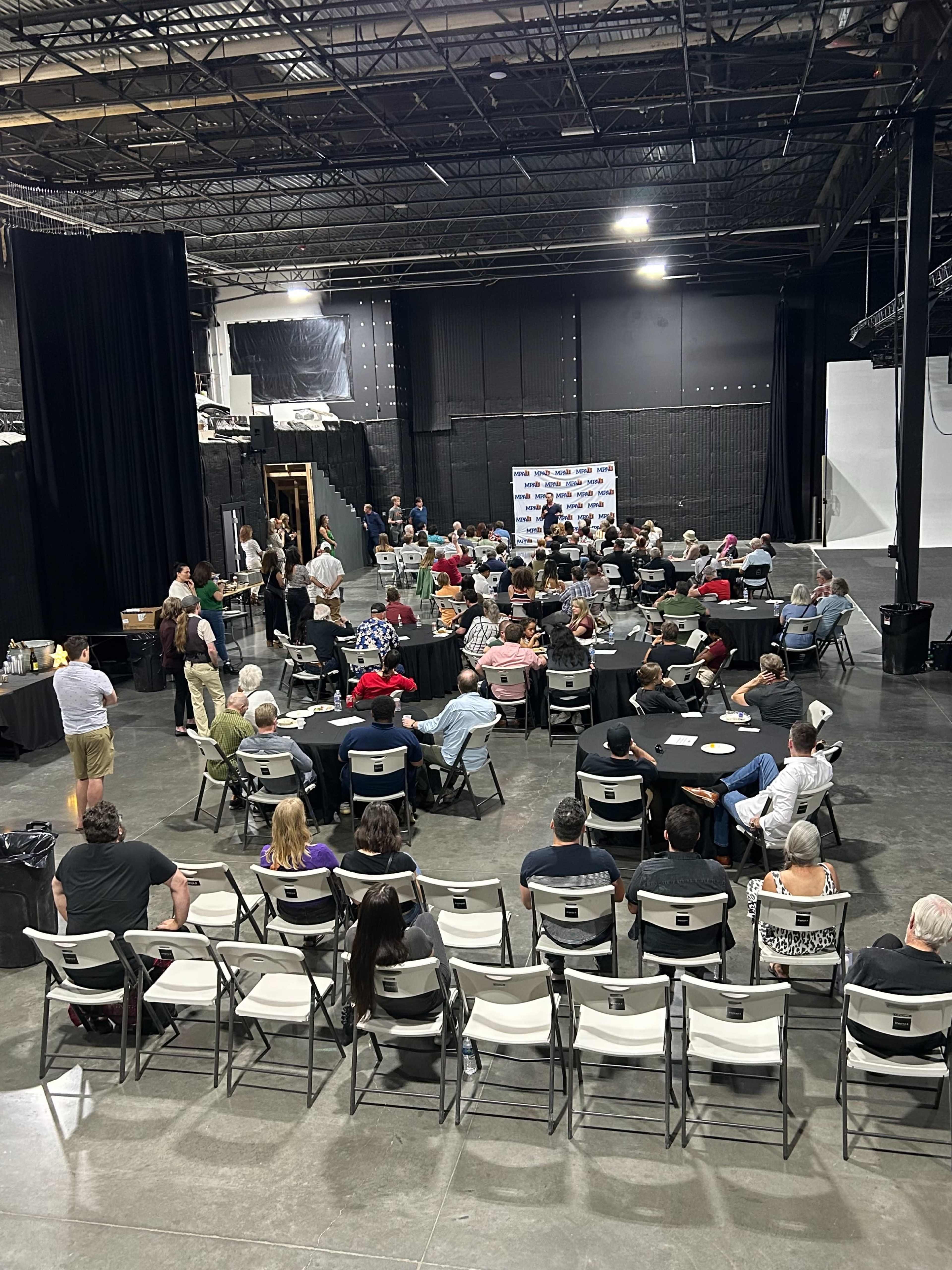 A large group of people is seated at round tables in a spacious indoor venue, with a stage and backdrop at one end.