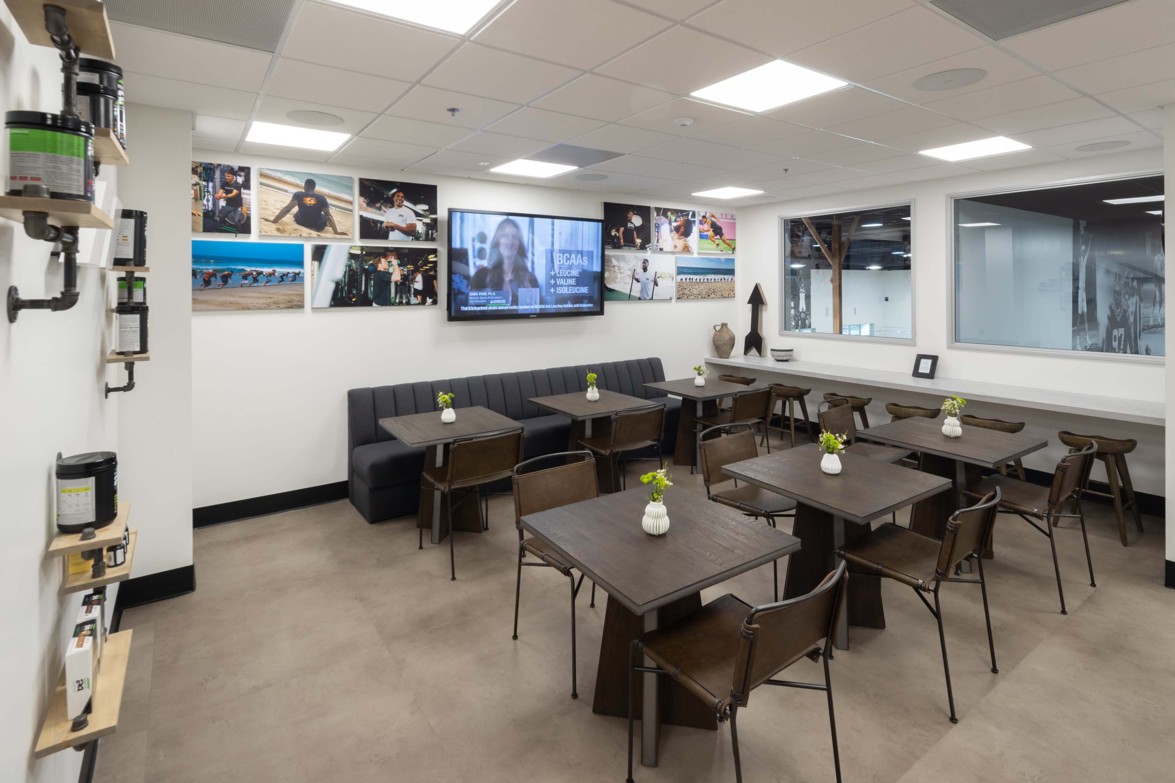 The image shows a modern dining area featuring several small tables with metal chairs, a wall-mounted television, and framed photos displayed on the walls.