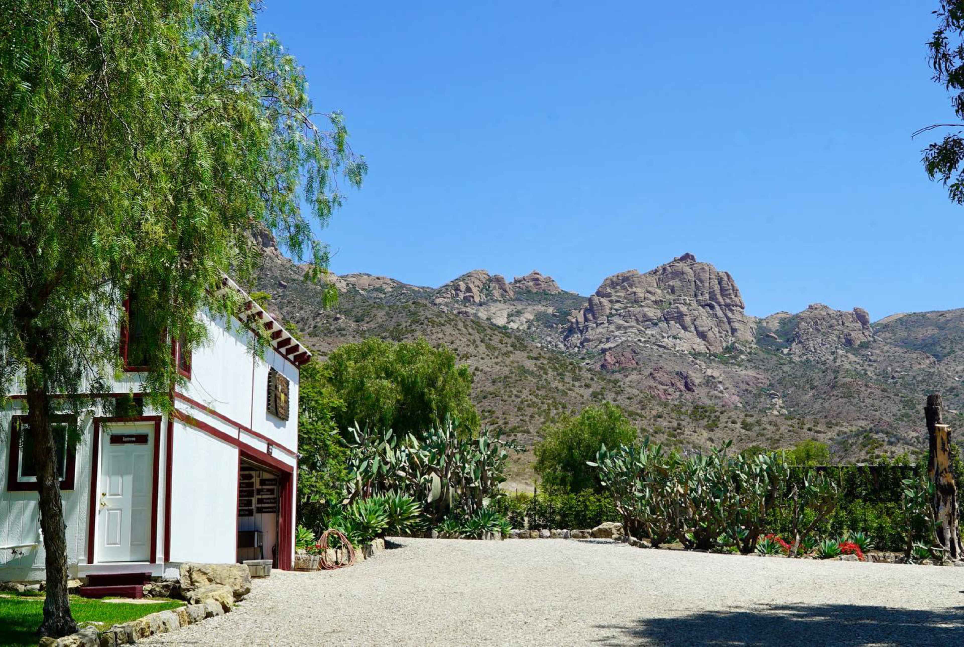 A small white building with a red roof is situated beside a gravel driveway, with rocky mountains and cacti in the background under a clear blue sky.