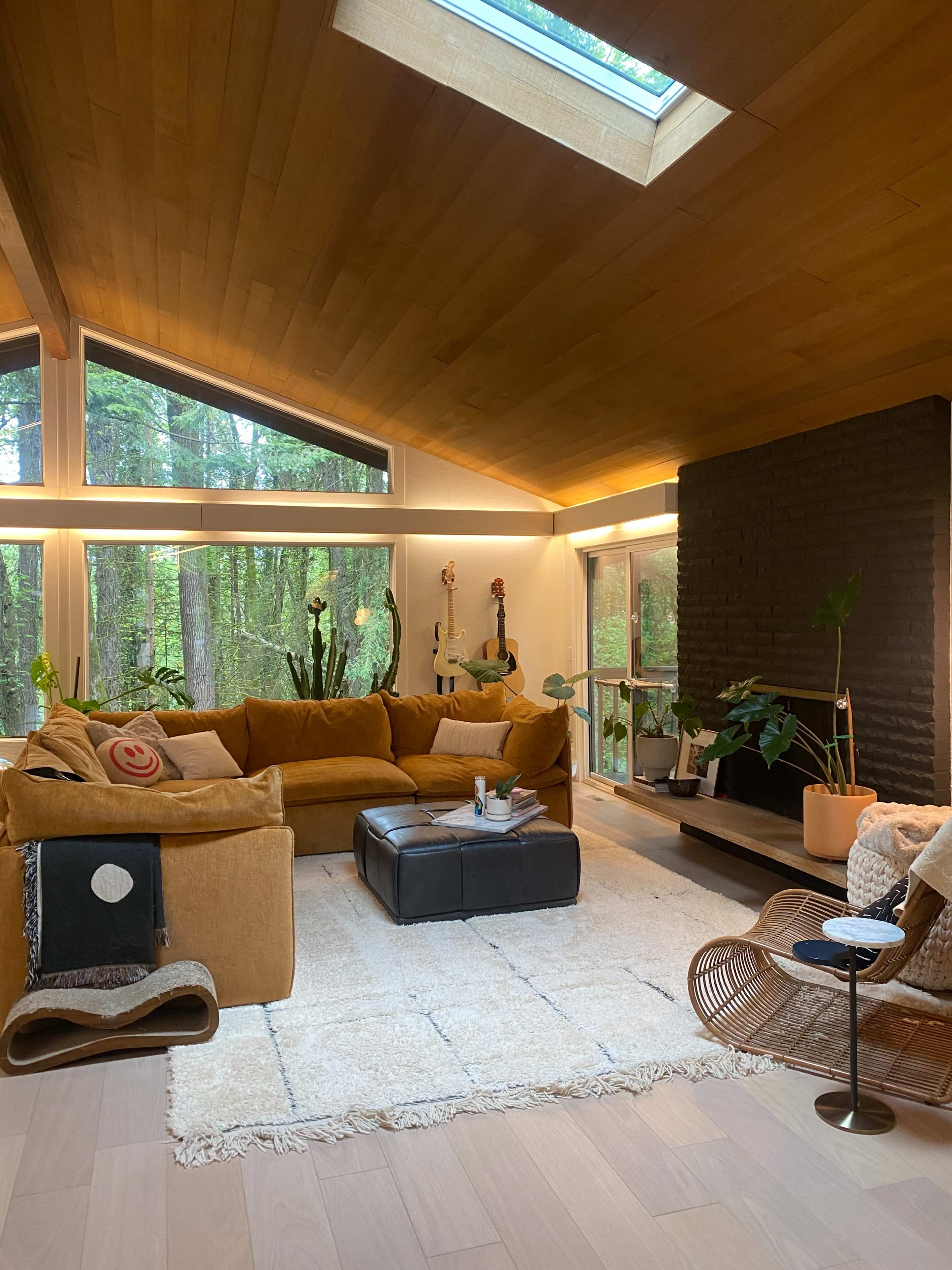 The living room features a large sectional sofa, a central ottoman, and a fireplace, all set against a backdrop of large windows overlooking a forest.