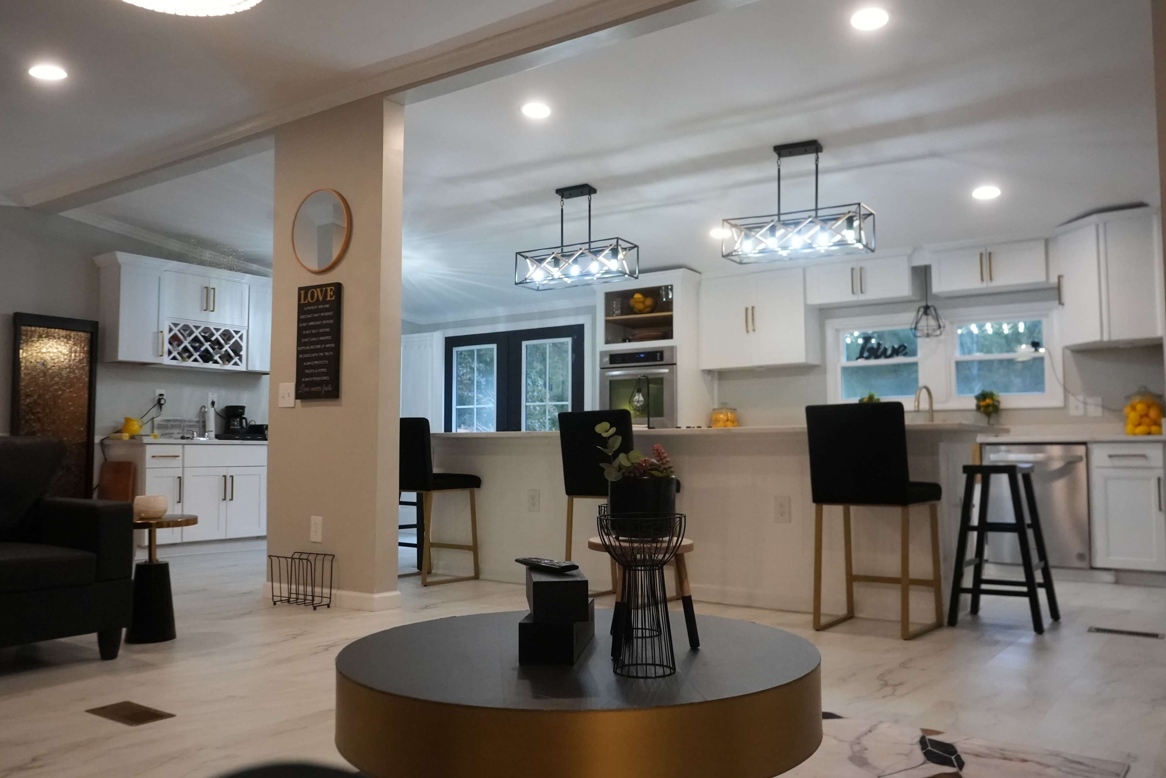 The image shows a modern kitchen and living area with white cabinetry, a large island with stools, and contemporary lighting fixtures.