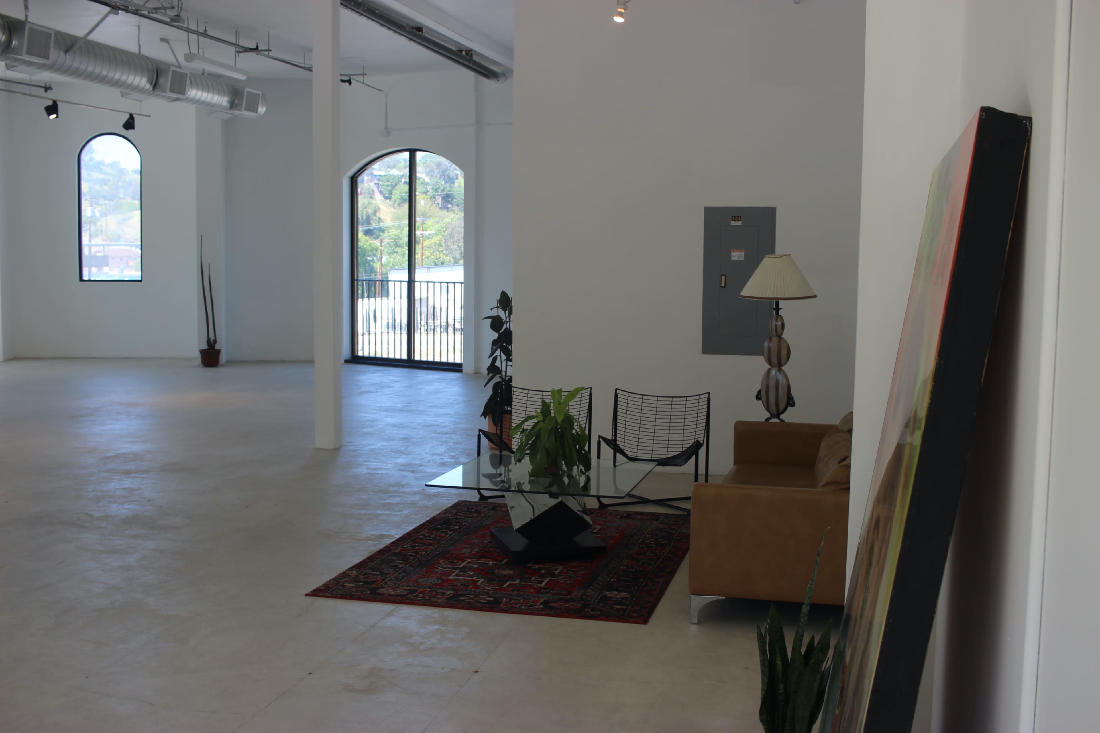 A spacious, minimalistic interior with a brown couch, a glass coffee table on a patterned rug, and large windows that overlook a green landscape.