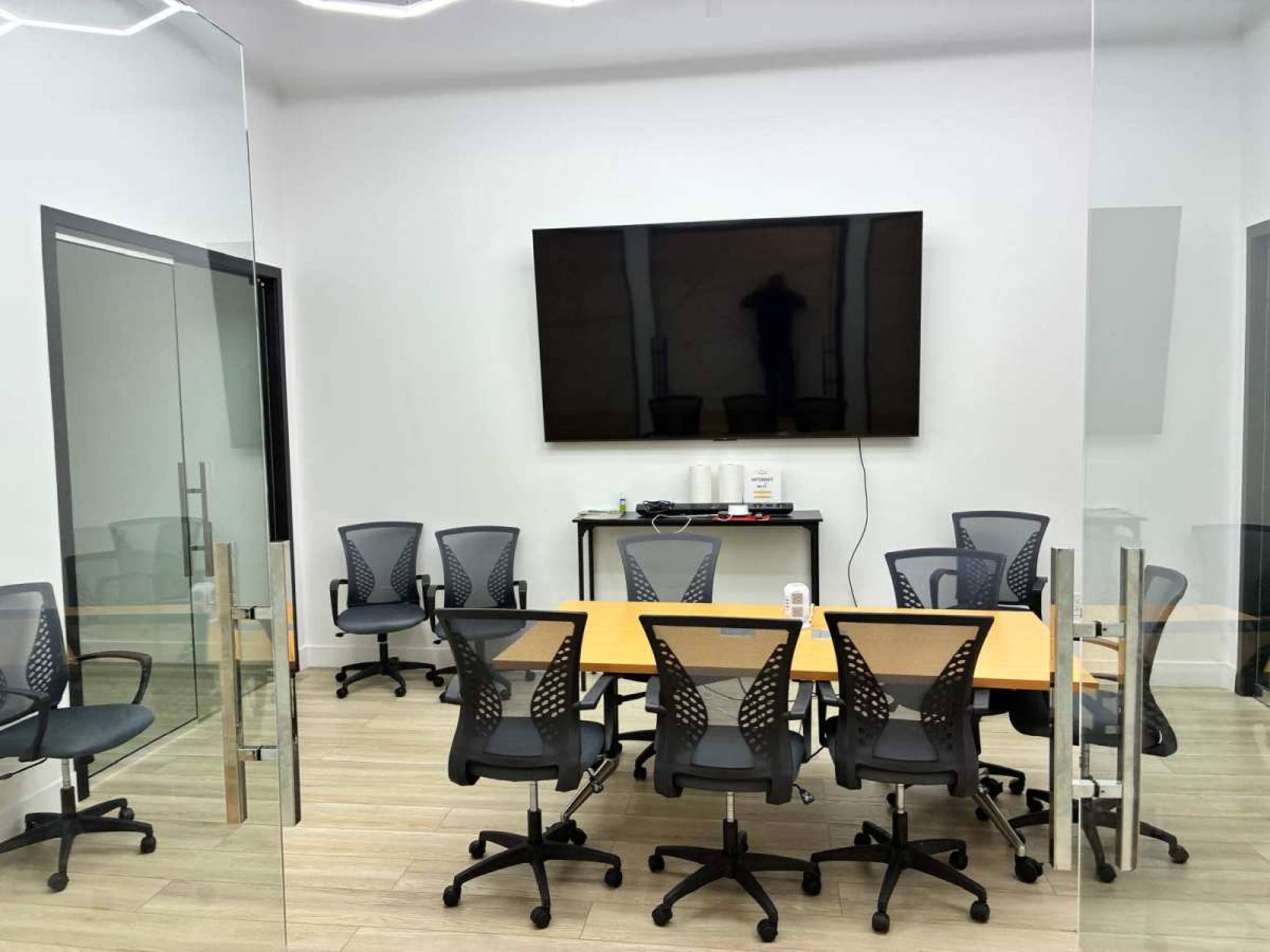 The image shows a modern conference room with a large wall-mounted screen, a central table surrounded by office chairs, and glass walls.
