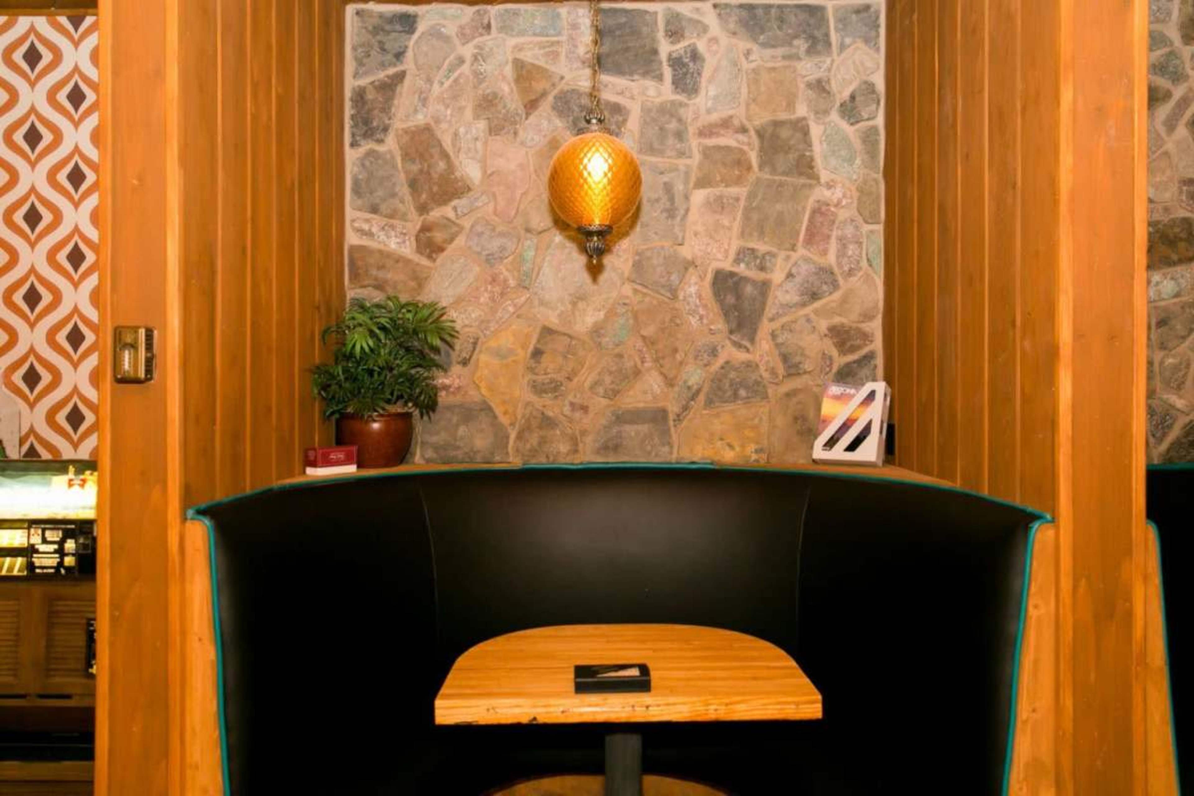 A black booth with a wooden table is set against a stone wall, illuminated by a pendant light.