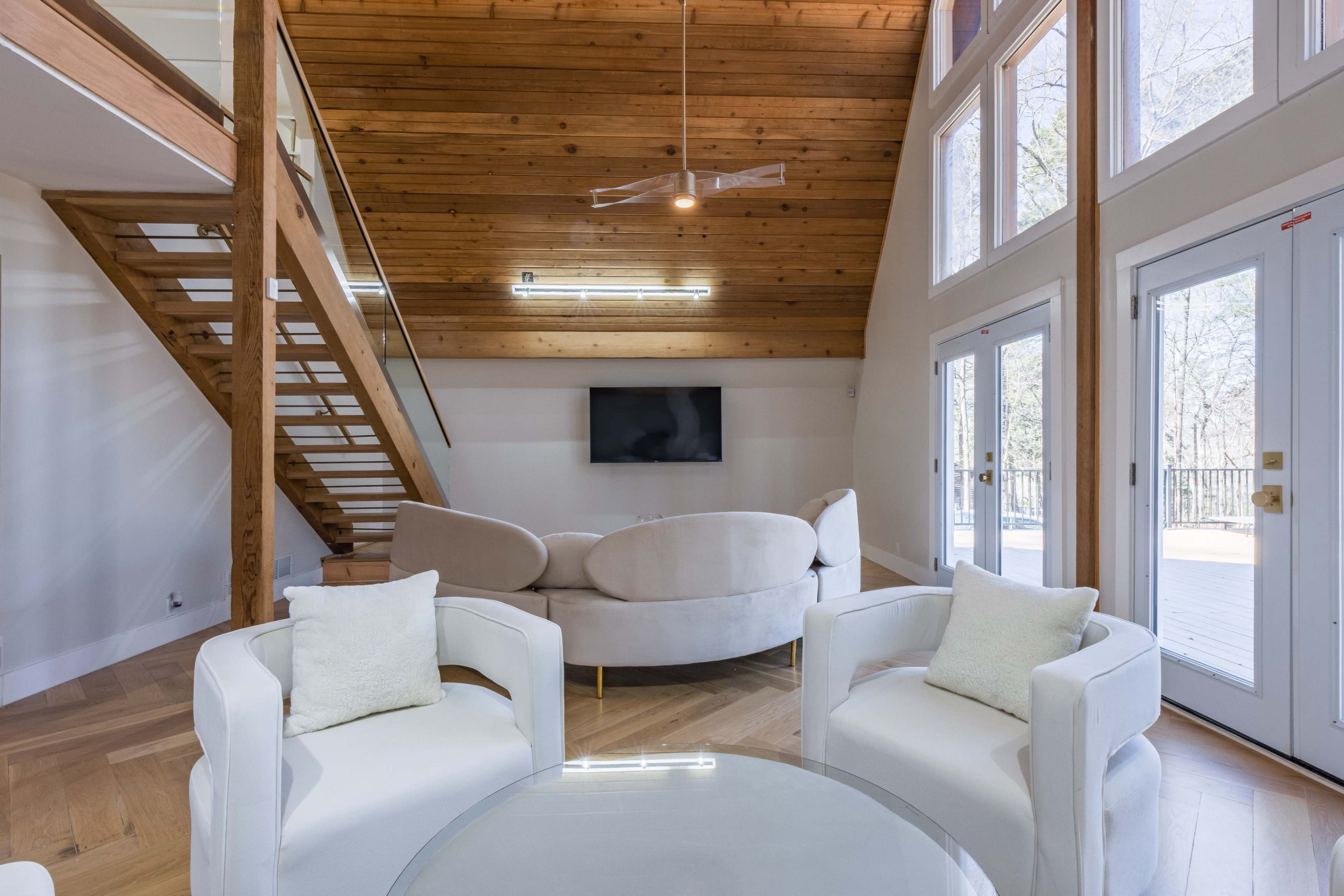 The interior features a modern living space with a wooden ceiling, a staircase, a circular sofa, and a glass table adjacent to large glass doors leading to an outdoor deck.