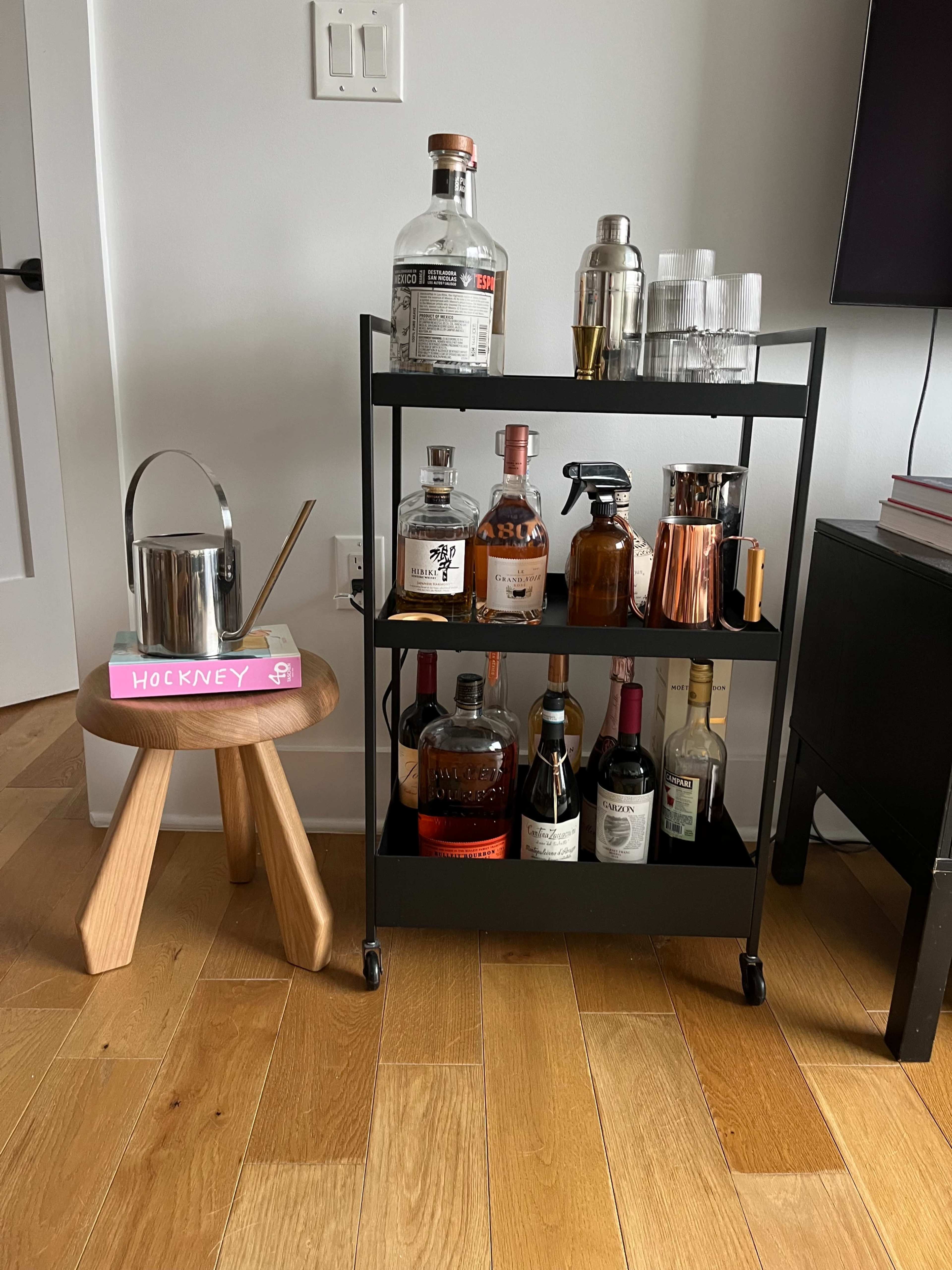A black bar cart with various bottles of liquor, a cocktail shaker, and glassware, positioned beside a wooden stool labeled "Hockney."