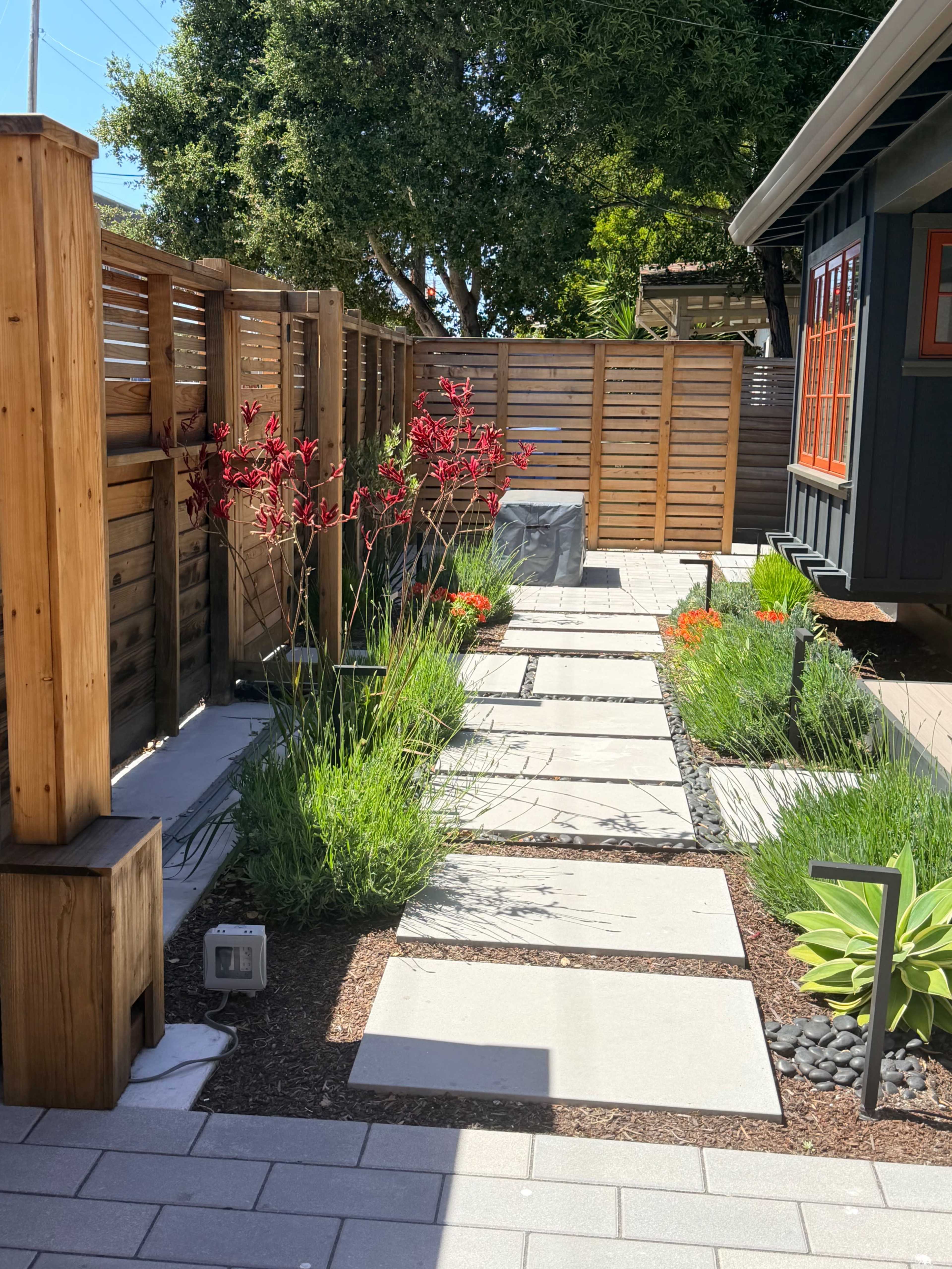 The image shows a landscaped pathway with concrete stepping stones surrounded by lush green plants and decorative red flowers, leading to a wooden fence and a house with orange window frames.