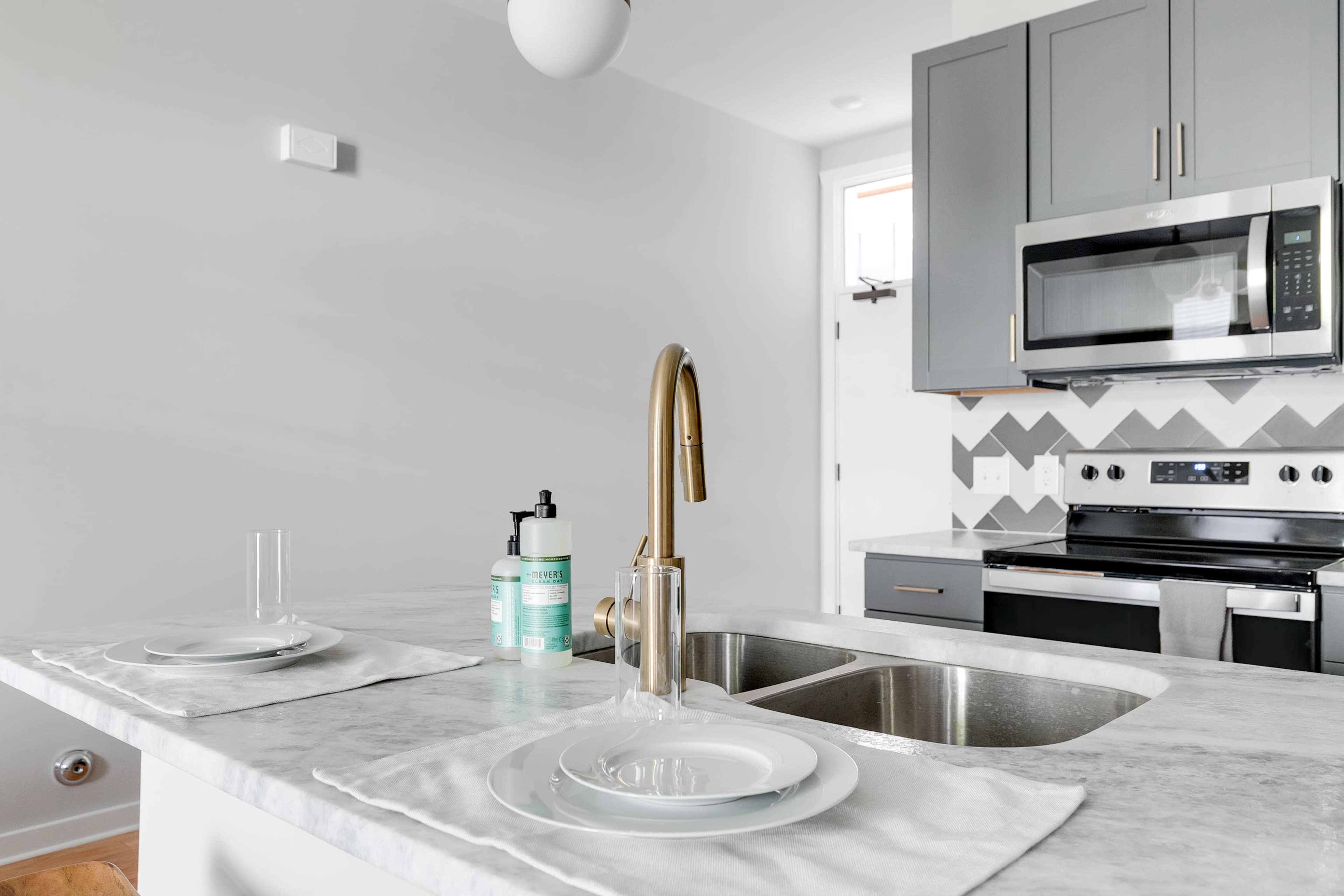 A modern kitchen with a marble countertop, stainless steel appliances, a double sink, and gray cabinets, complemented by chevron patterned tiles on the backsplash.
