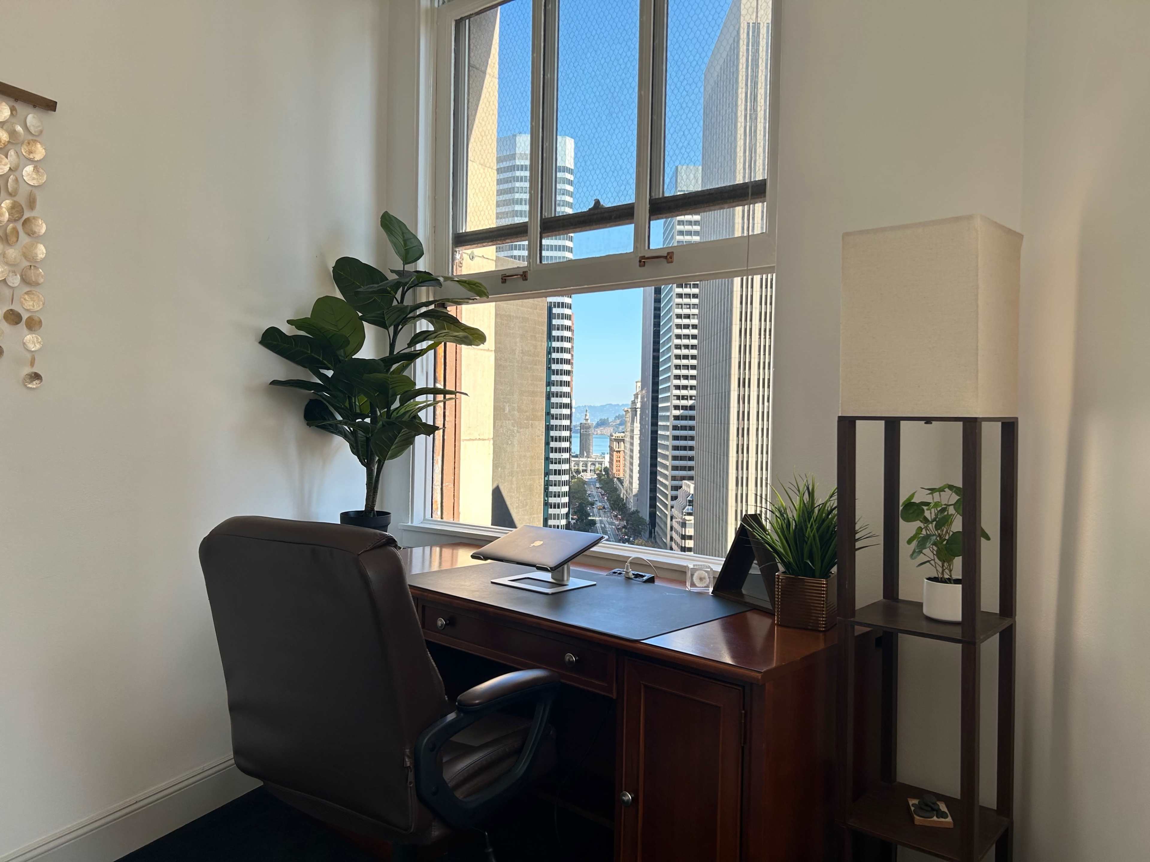 A wooden desk with a chair, a laptop, and small plants sits beside a large window overlooking tall buildings and a distant view of water.