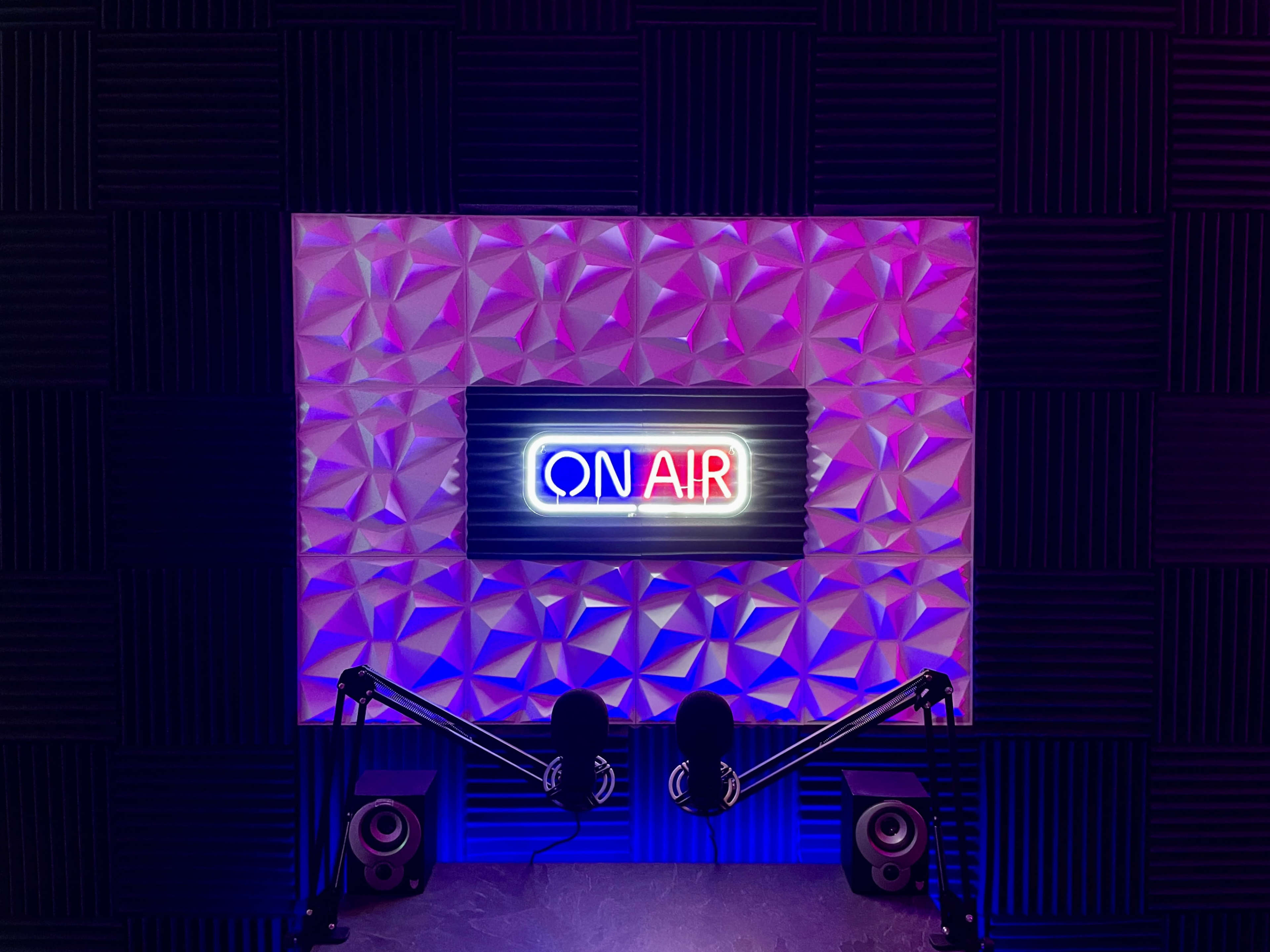 A studio setup featuring a "ON AIR" neon sign against a textured wall, with two microphones and speakers positioned below it.