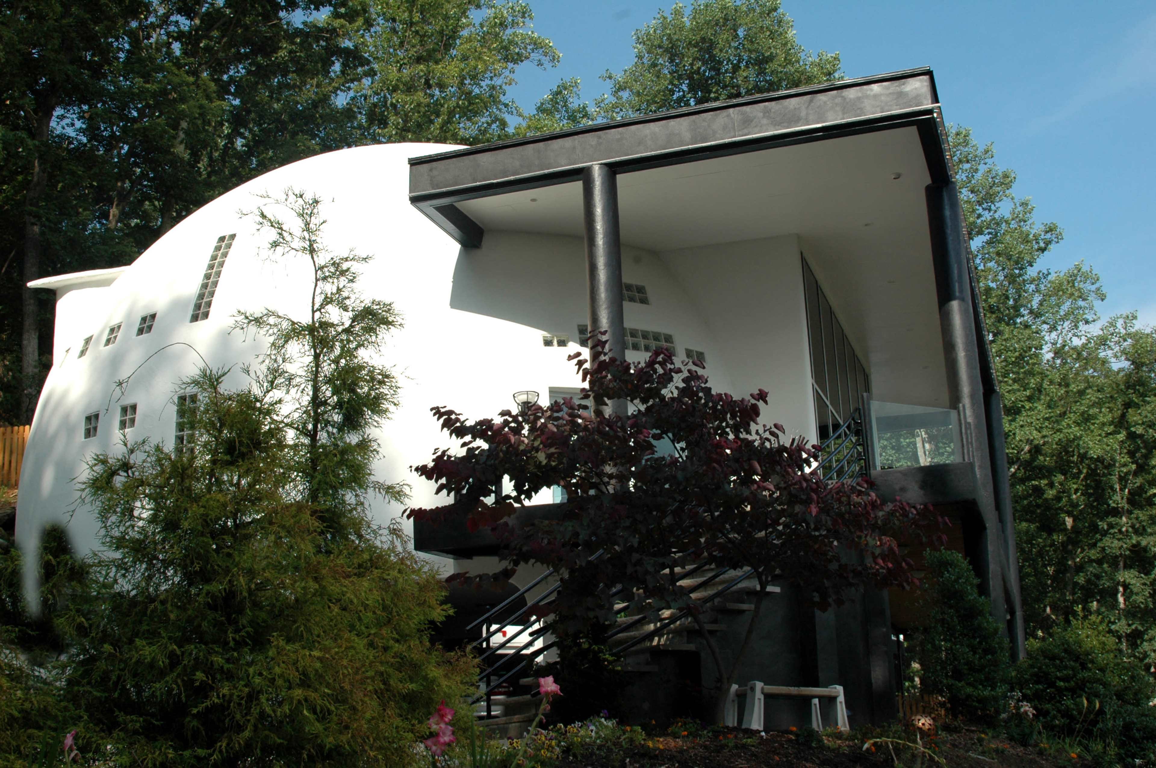 A modern, rounded white house with large glass windows and a black overhanging roof, surrounded by greenery.