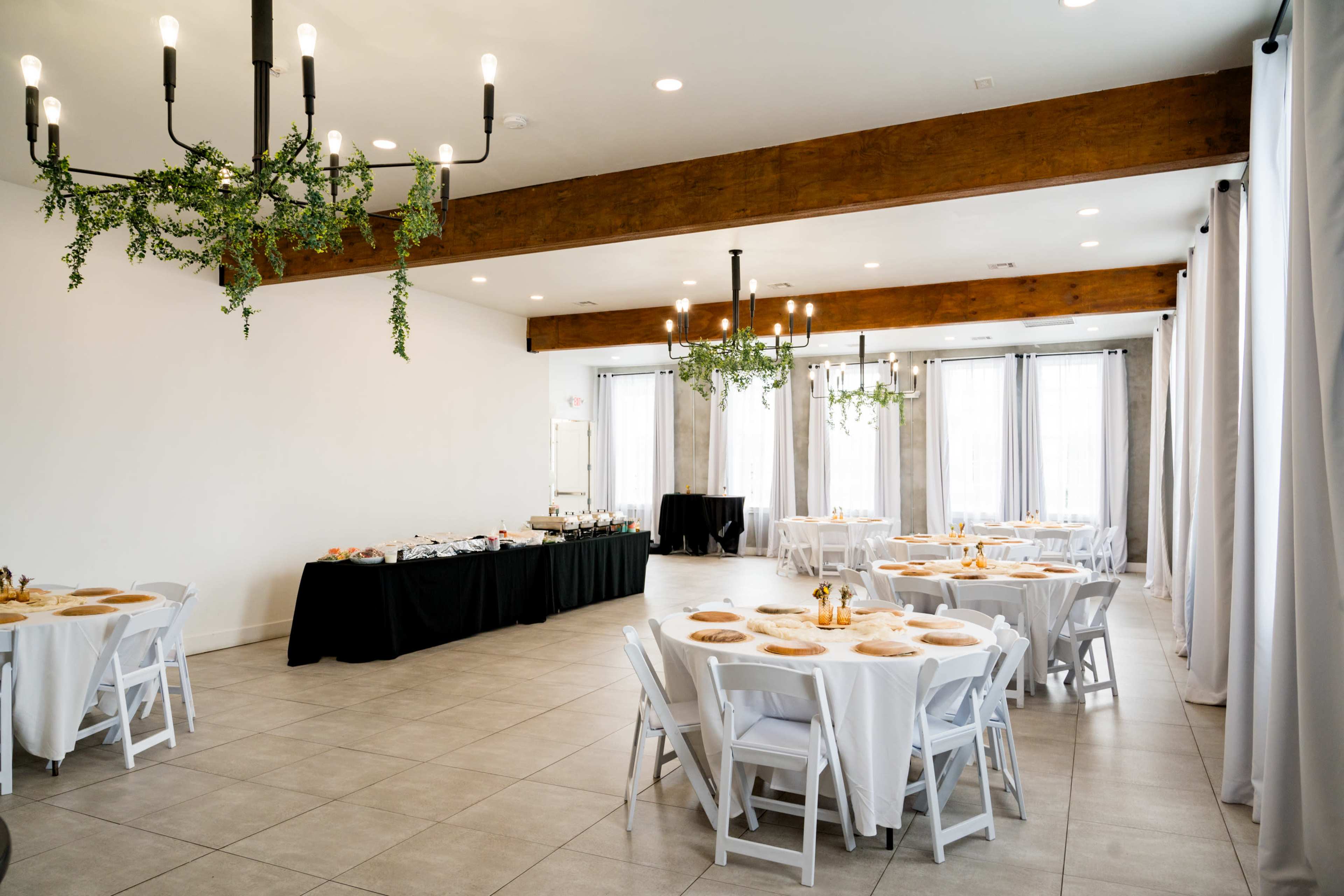 A well-lit event space features round tables with white tablecloths and chairs, complemented by greenery hanging from modern light fixtures.