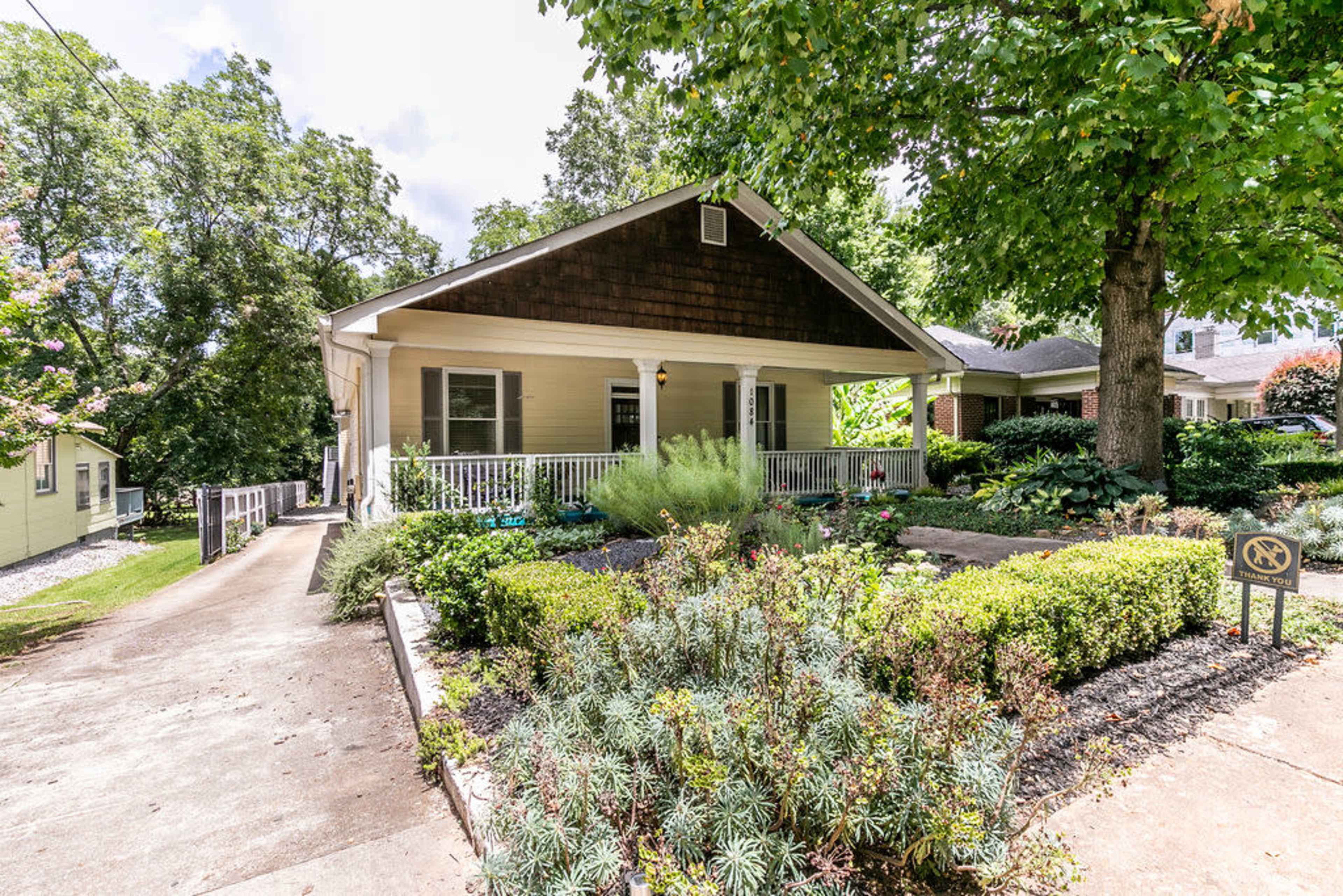 A single-story house with a front porch is surrounded by a landscaped yard featuring various plants and shrubs.
