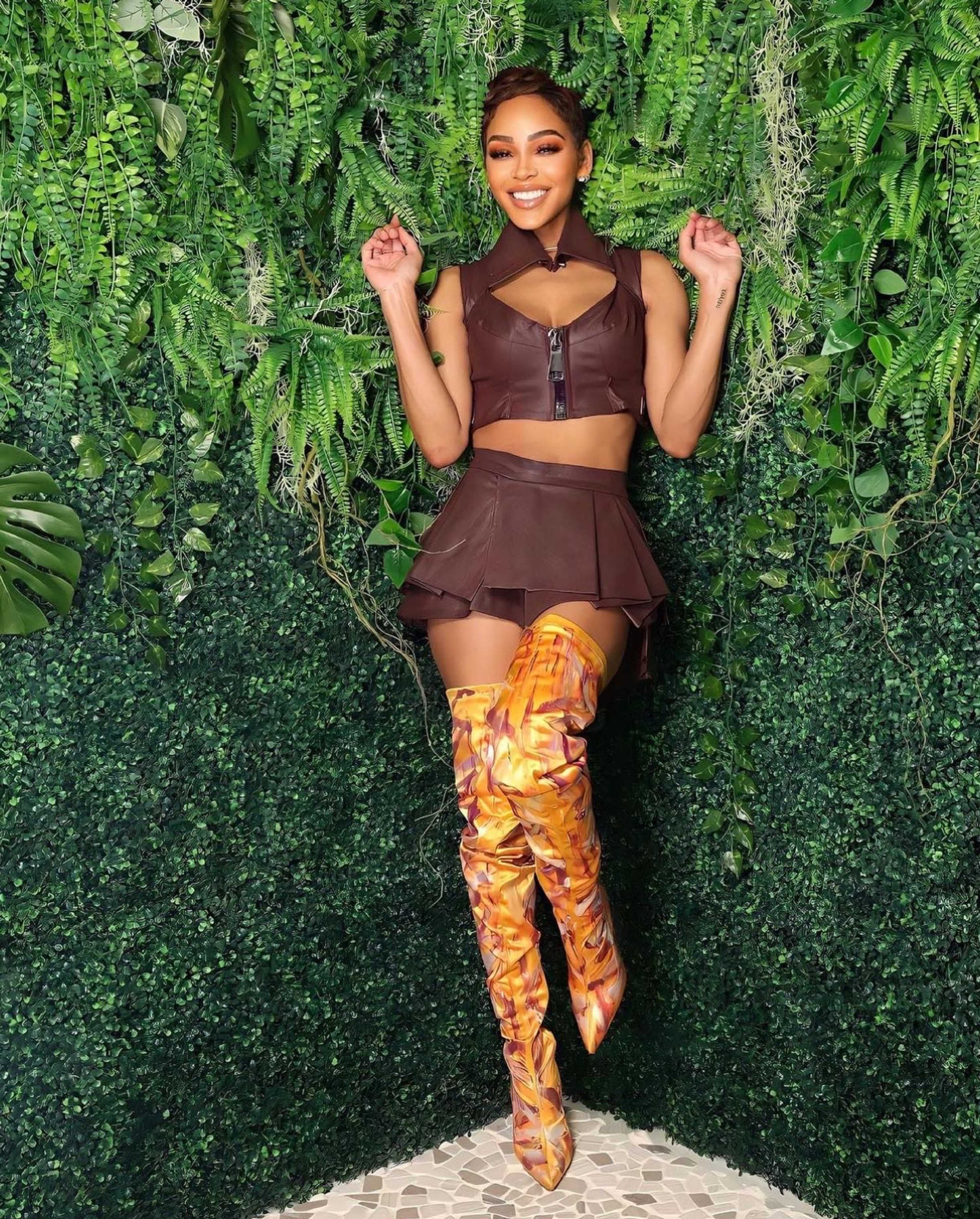 A woman poses against a green wall adorned with plants, wearing a brown cropped top and a matching skirt, along with knee-high, brightly colored boots.