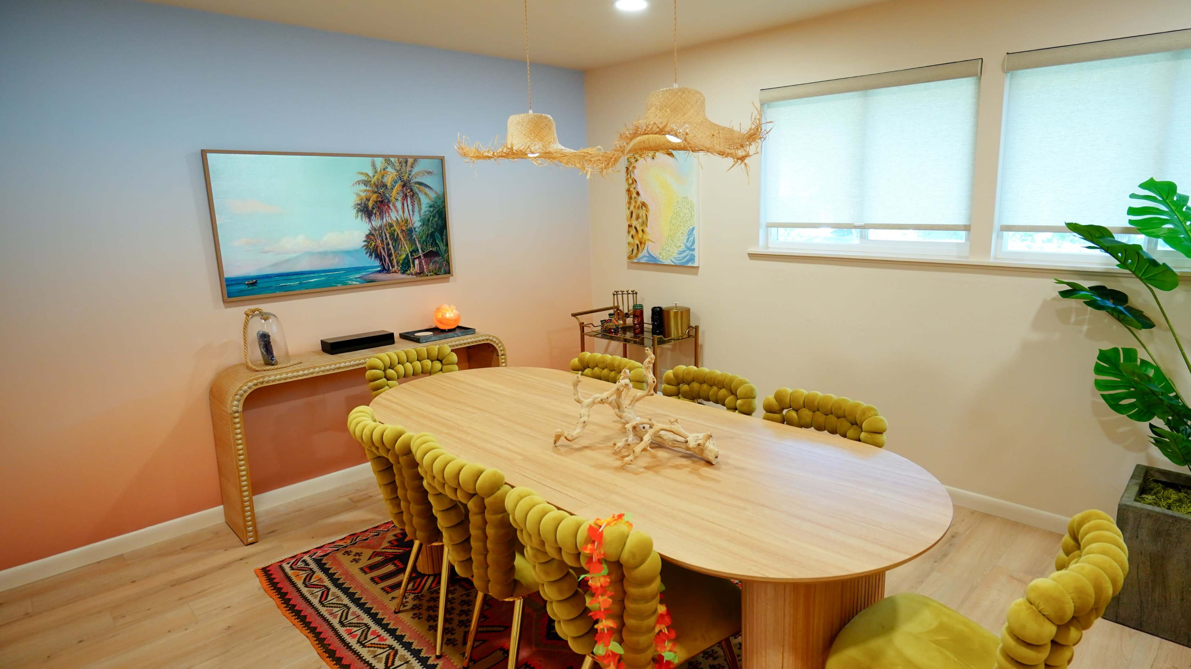 The image shows a modern dining room with a large oval wooden table surrounded by green upholstered chairs, a wall-mounted painting of a beach scene, and a decorative pendant light.