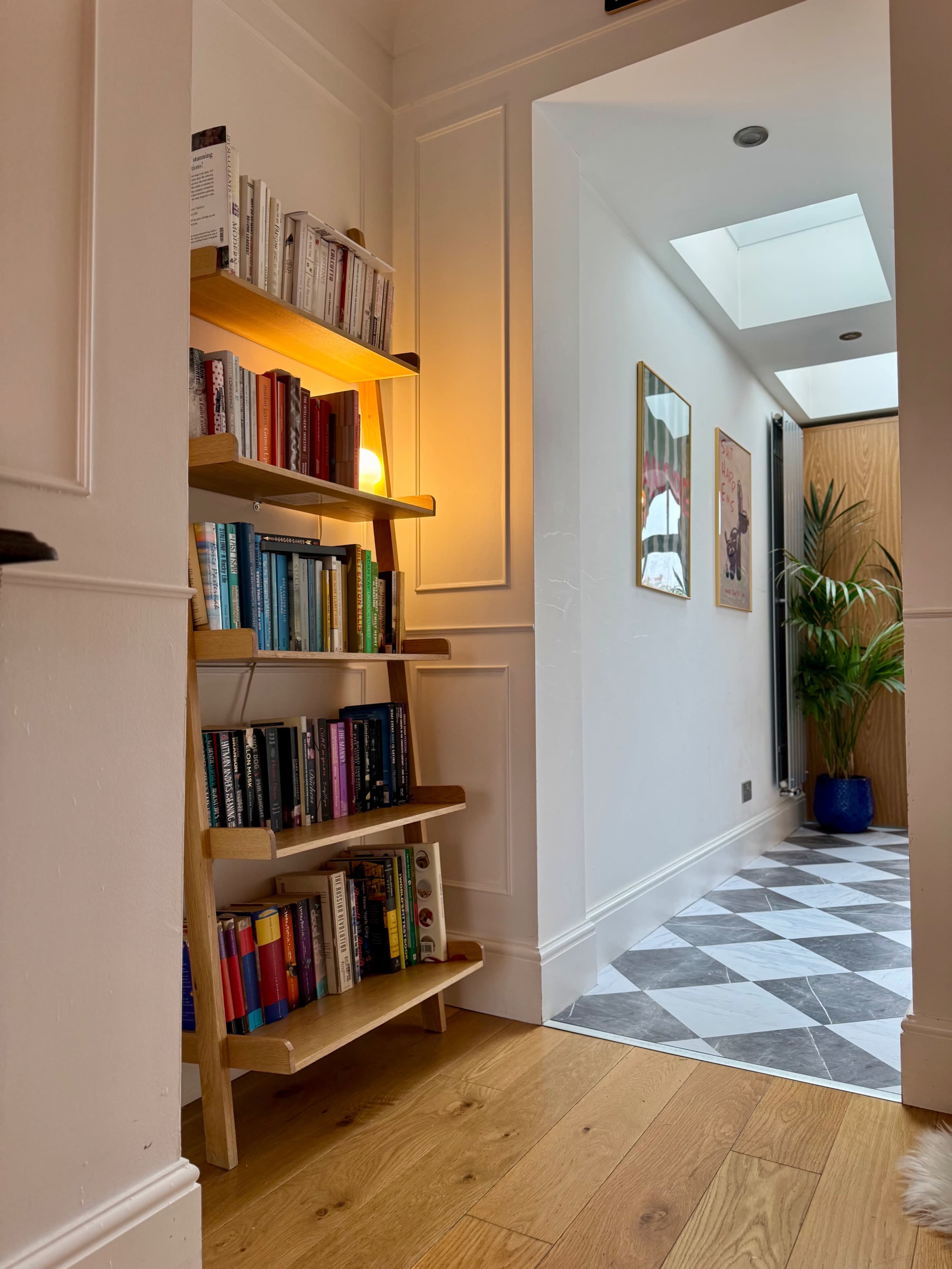 A wooden bookshelf with colorful books stands in a hallway that leads to a room with a skylight and decorative artwork on the walls.