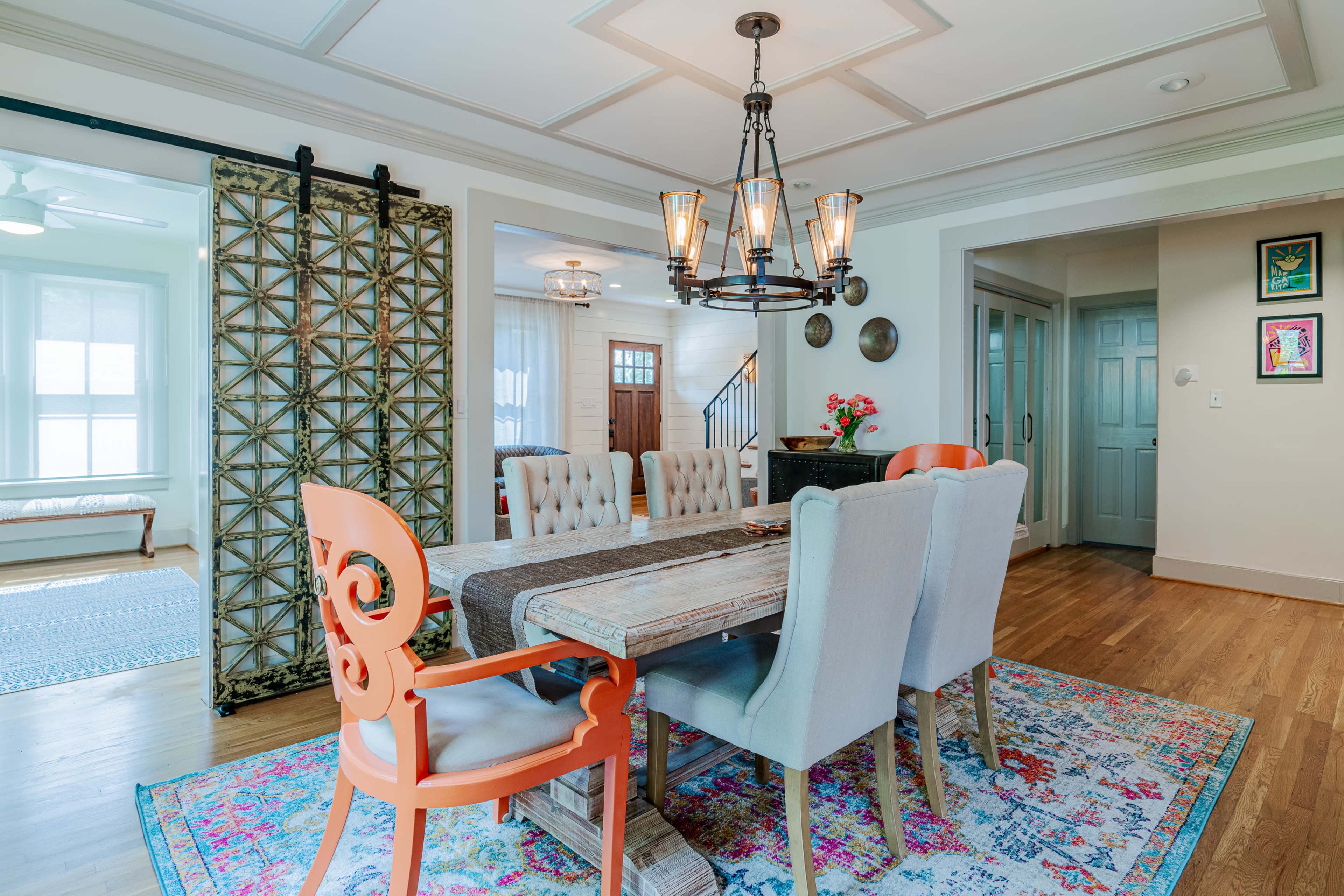 A dining area with a wooden table surrounded by upholstered chairs, a decorative chandelier overhead, and a patterned area rug on the hardwood floor.
