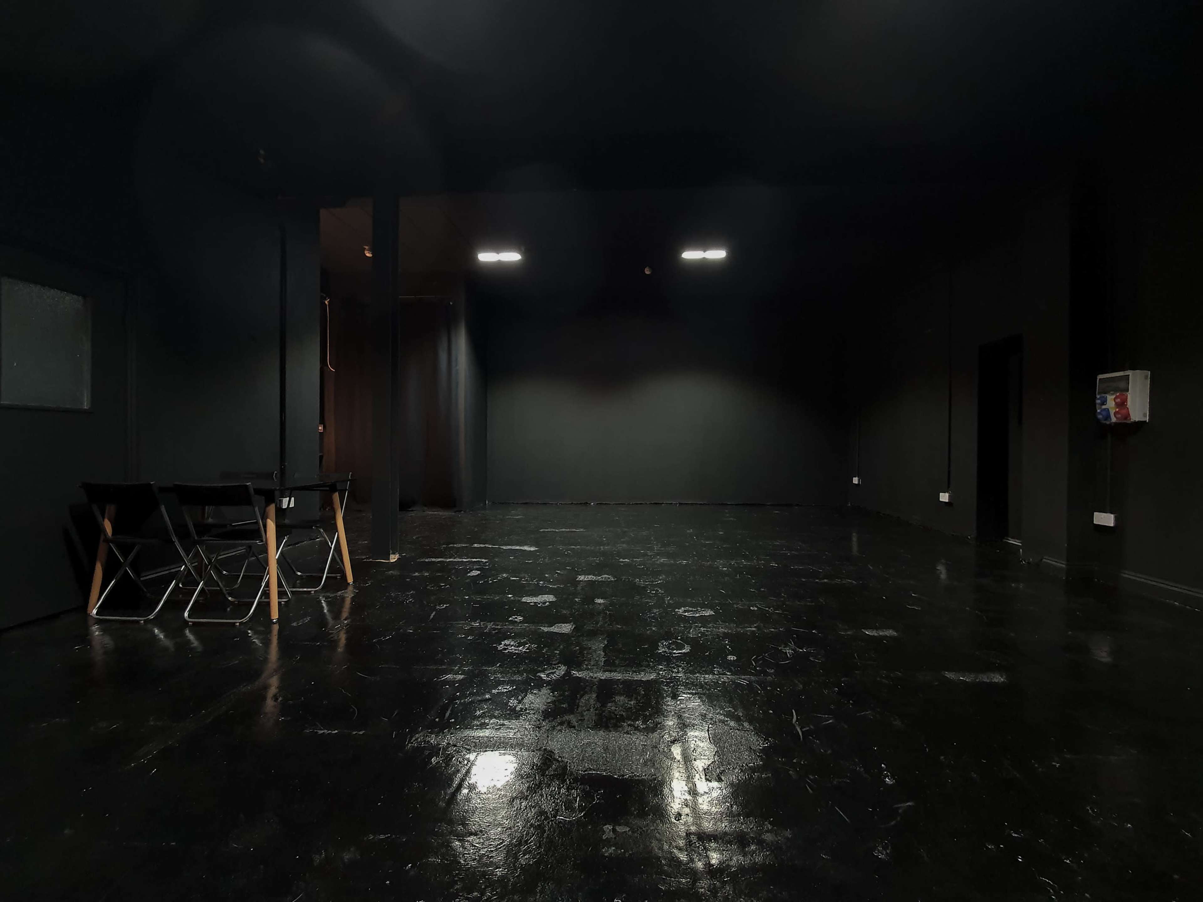 A large, empty room with black walls and a shiny black floor, featuring a few folding chairs in one corner.