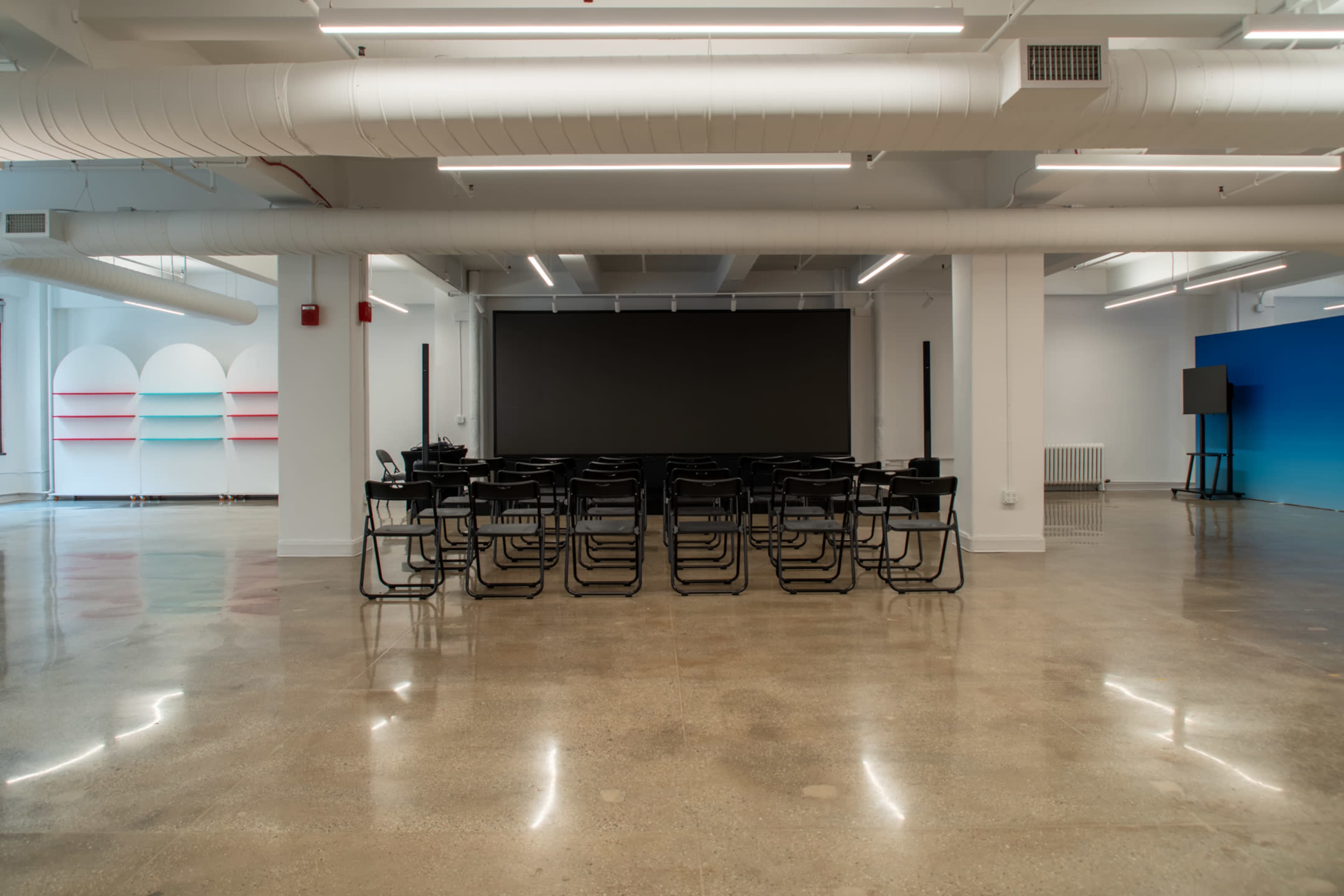 The image shows a spacious meeting room with rows of black chairs facing a large blank screen, complemented by colorful shelves against one wall.