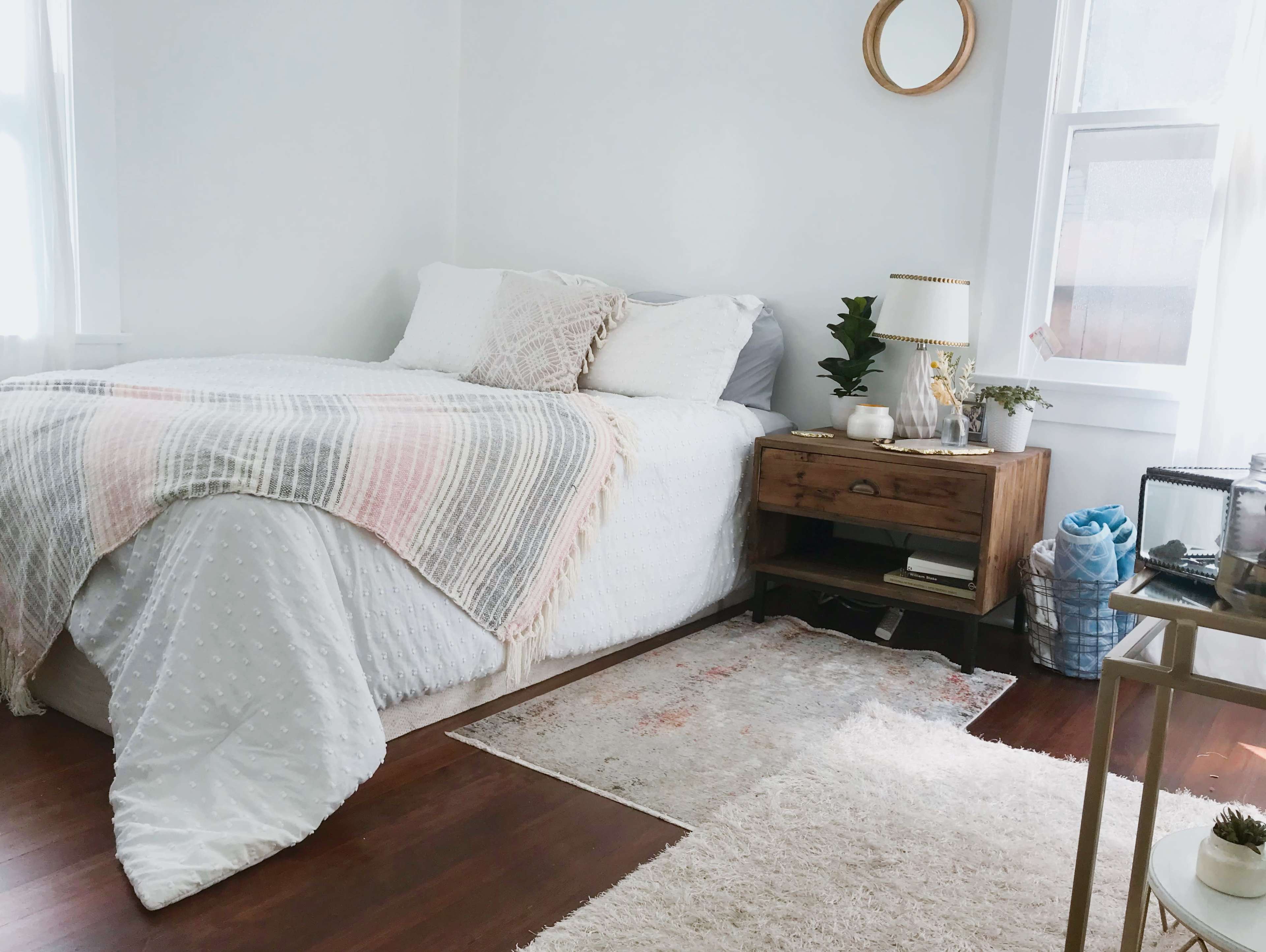 A neatly arranged bedroom features a bed with a patterned blanket, a wooden bedside table with plants and decor, and a light-colored area rug on the floor.