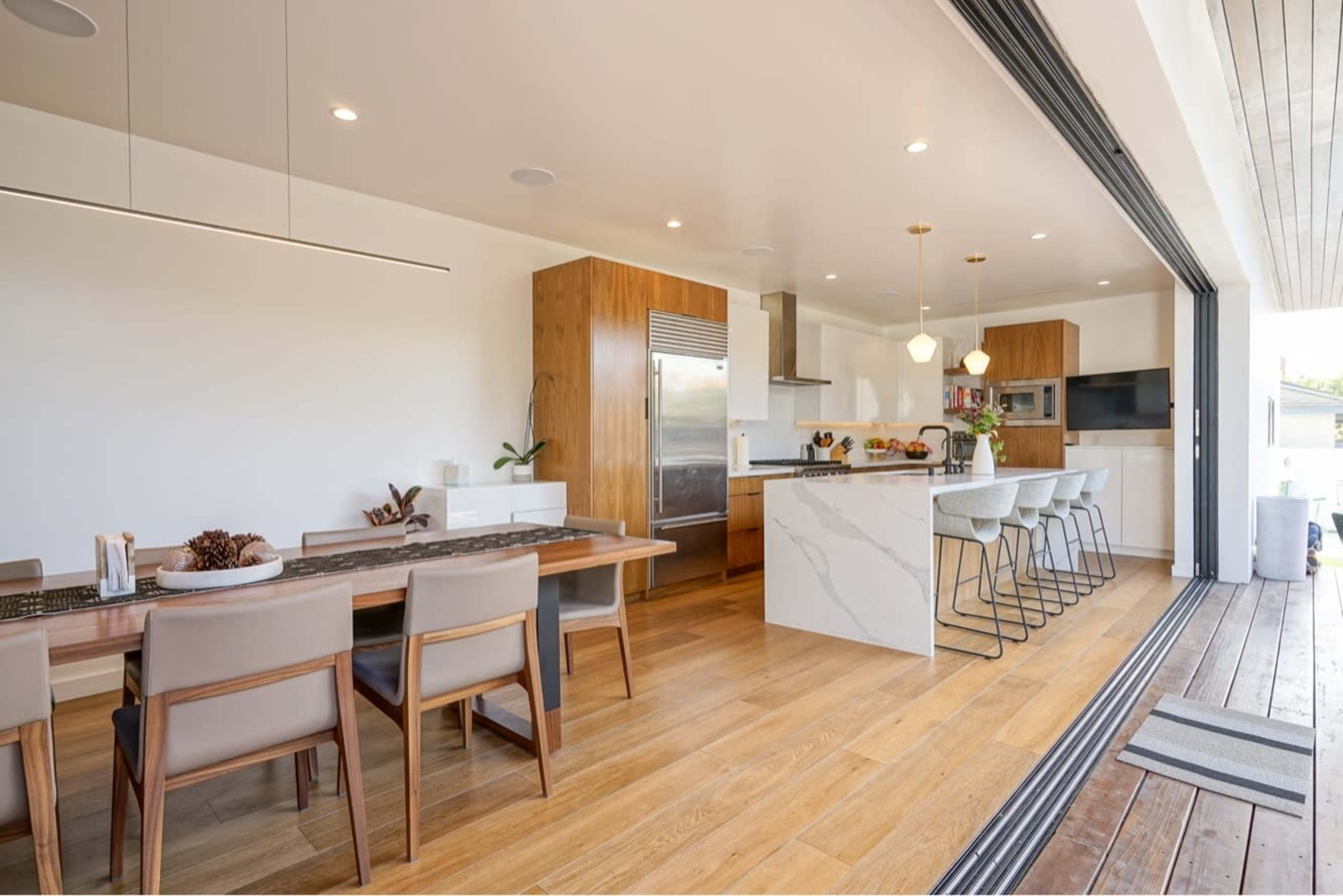 The image shows a modern kitchen and dining area featuring a large island, sleek cabinetry, and a dining table with chairs, all set in a spacious layout with an open design that connects to an outdoor space.