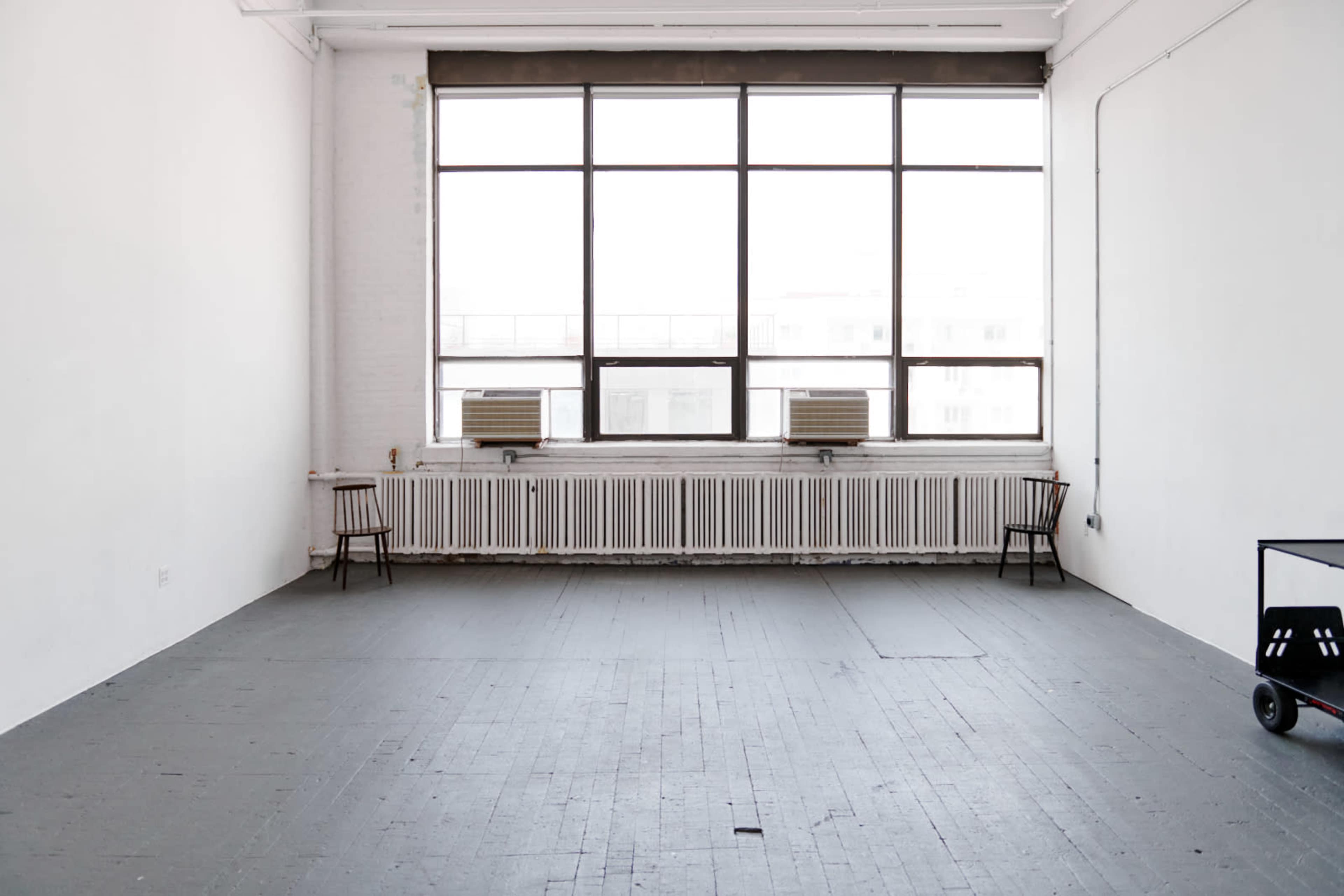 A spacious, empty room with two chairs and a large window, featuring air conditioning units and a radiator along one wall.
