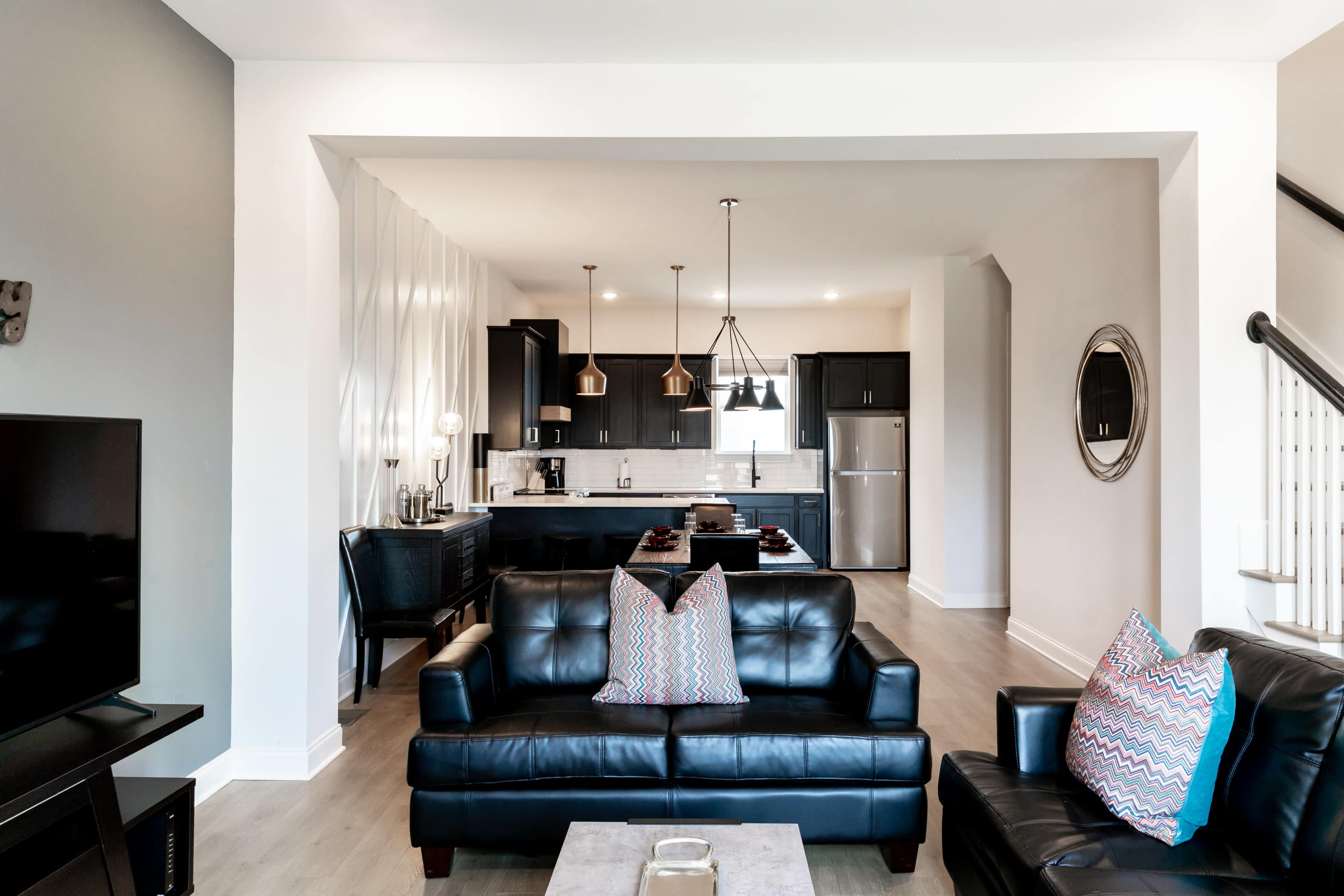 The image shows a modern living room with black leather sofas facing a kitchen area featuring dark cabinets and stainless steel appliances.
