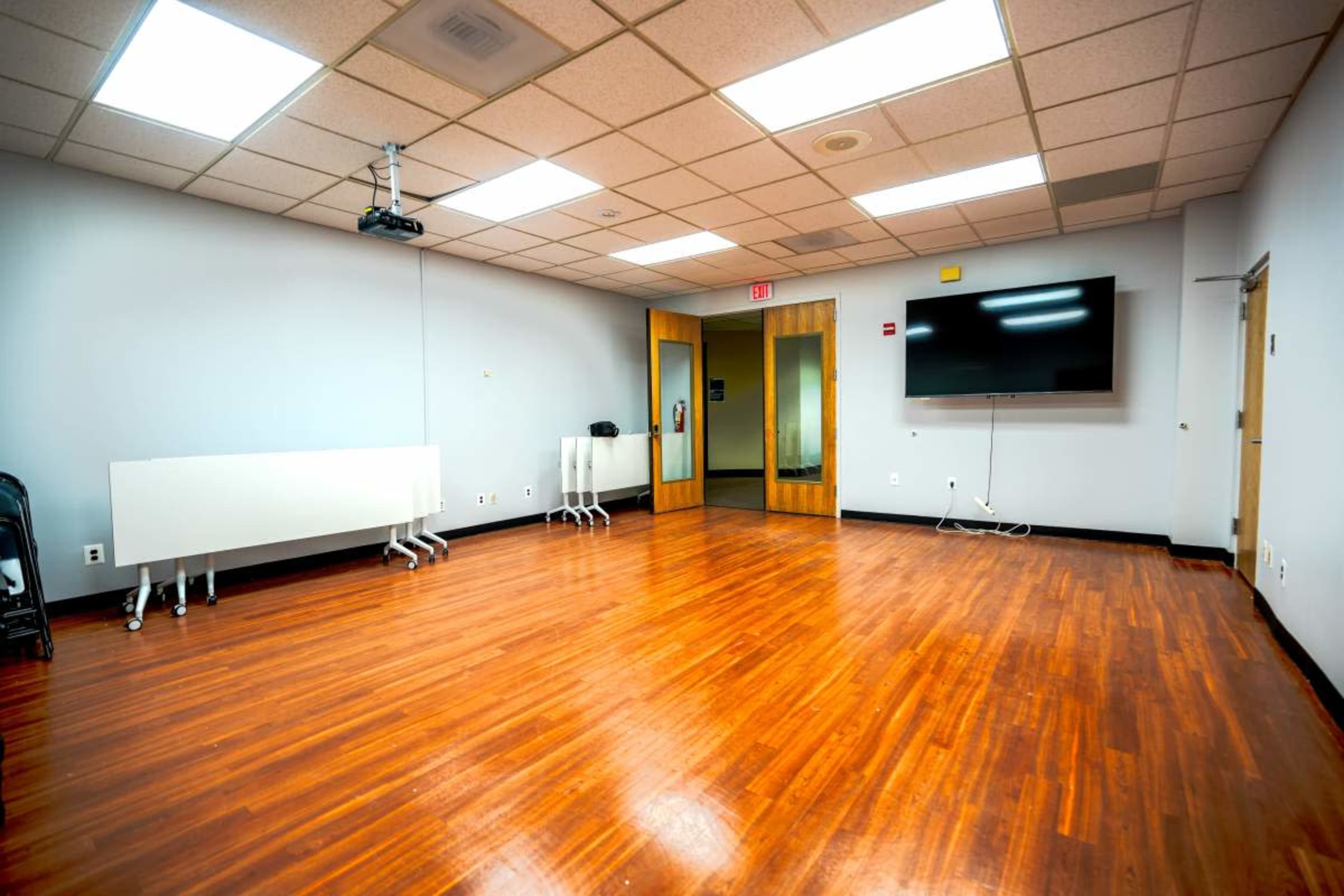 The image shows a spacious room with wooden flooring, equipped with a wall-mounted television and empty mobile whiteboards along one side.