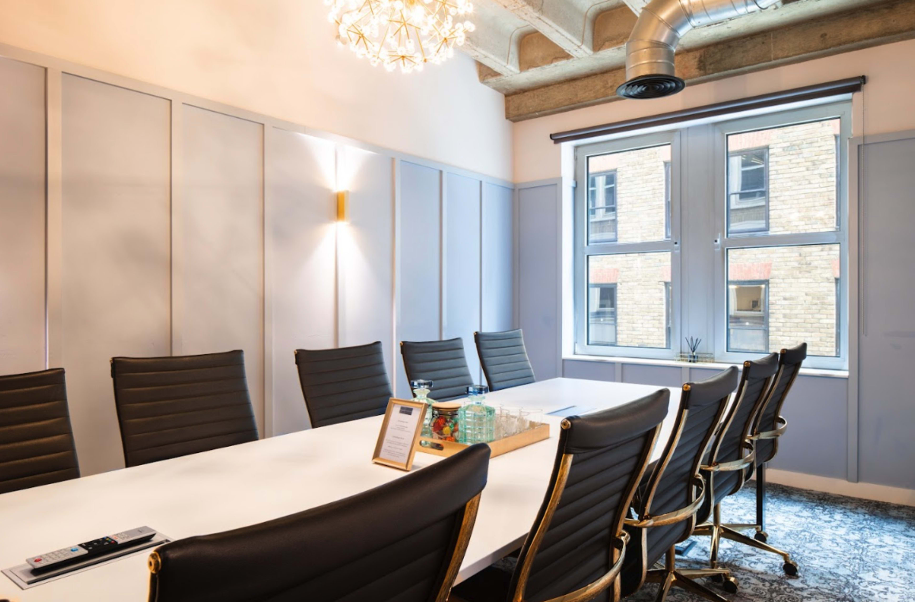 A conference room features a long white table surrounded by black leather chairs, illuminated by overhead lighting and natural light from large windows.