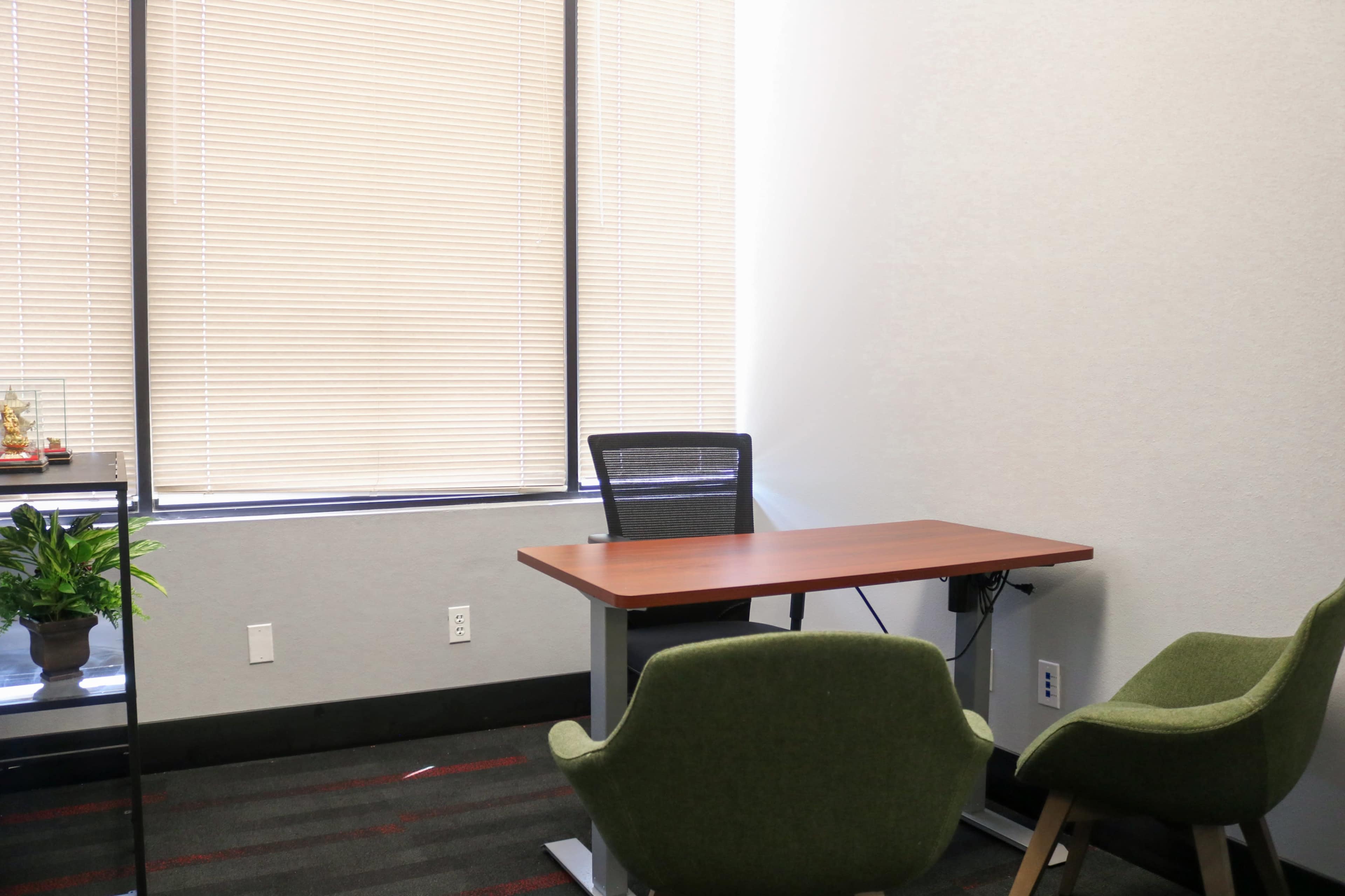 The image shows a small office space featuring a desk, a chair, and two green chairs, with a window partially covered by blinds.