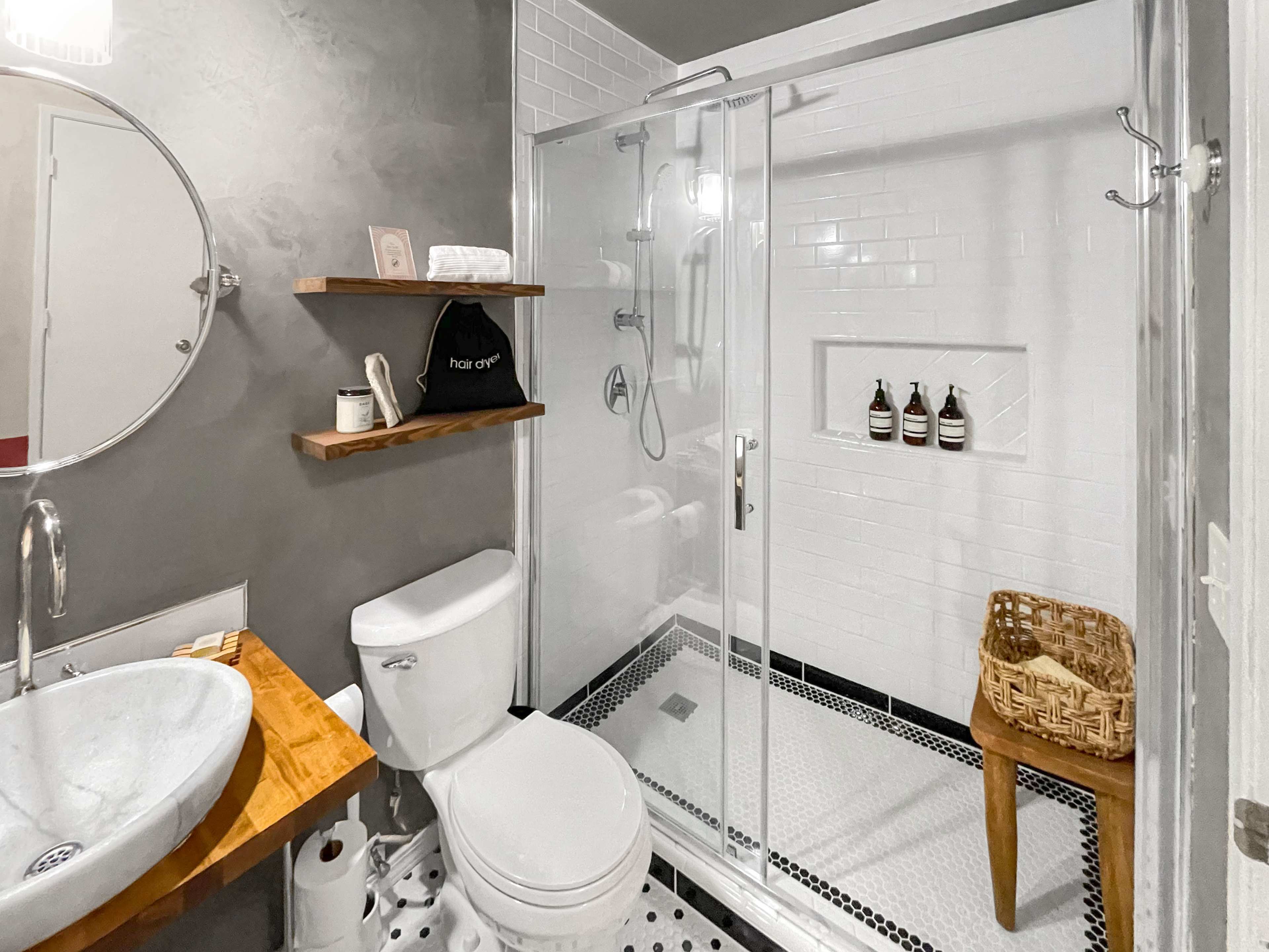 A modern bathroom features a glass shower enclosure, a circular mirror, a wooden sink counter, and a toilet, with decorative shelves holding toiletries.
