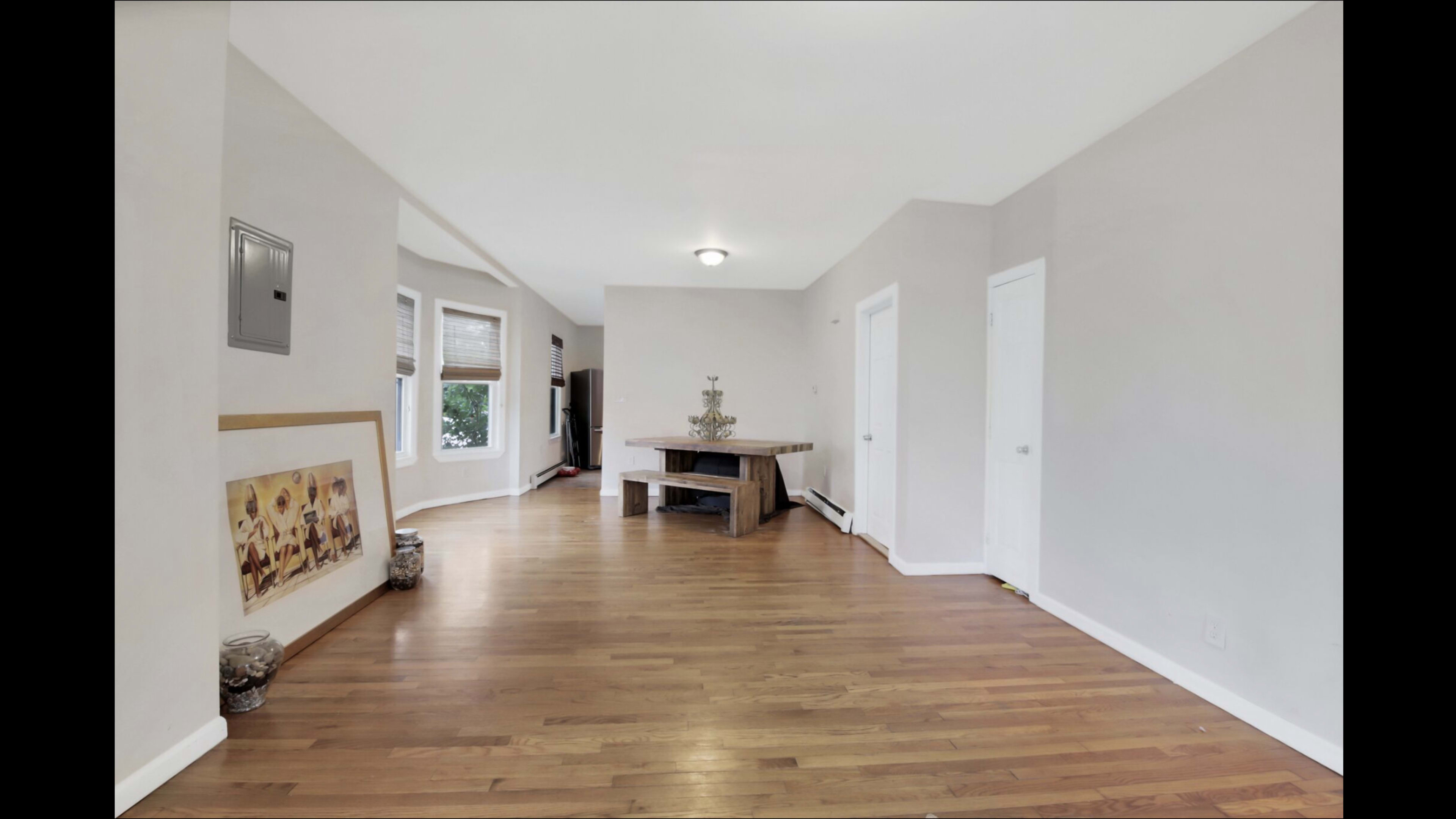 The image shows a spacious, light-filled interior room with hardwood flooring, a wooden table in the center, and doors leading to other areas.