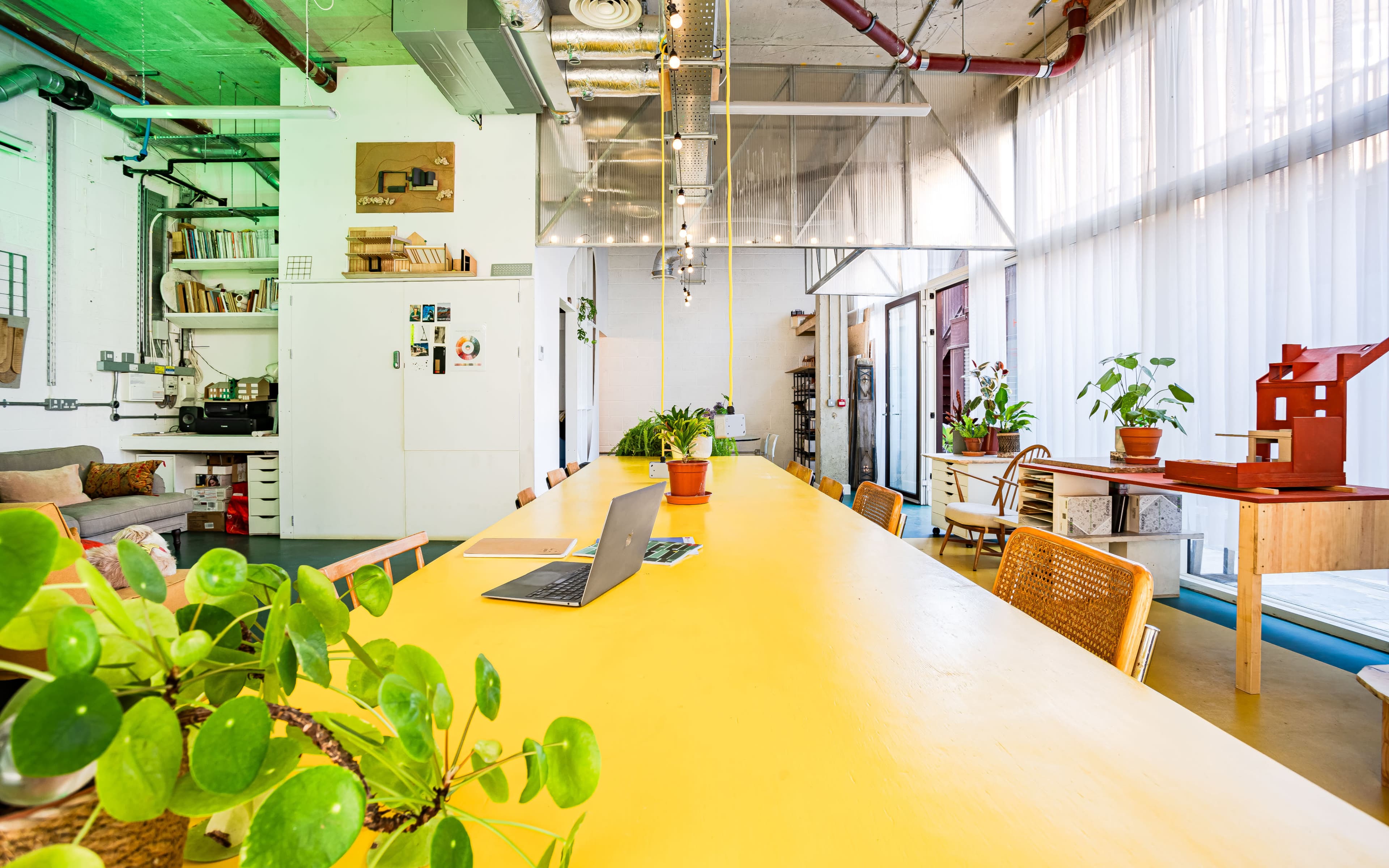 The image shows a brightly lit, modern workspace featuring a long yellow table, potted plants, and a variety of seating options.