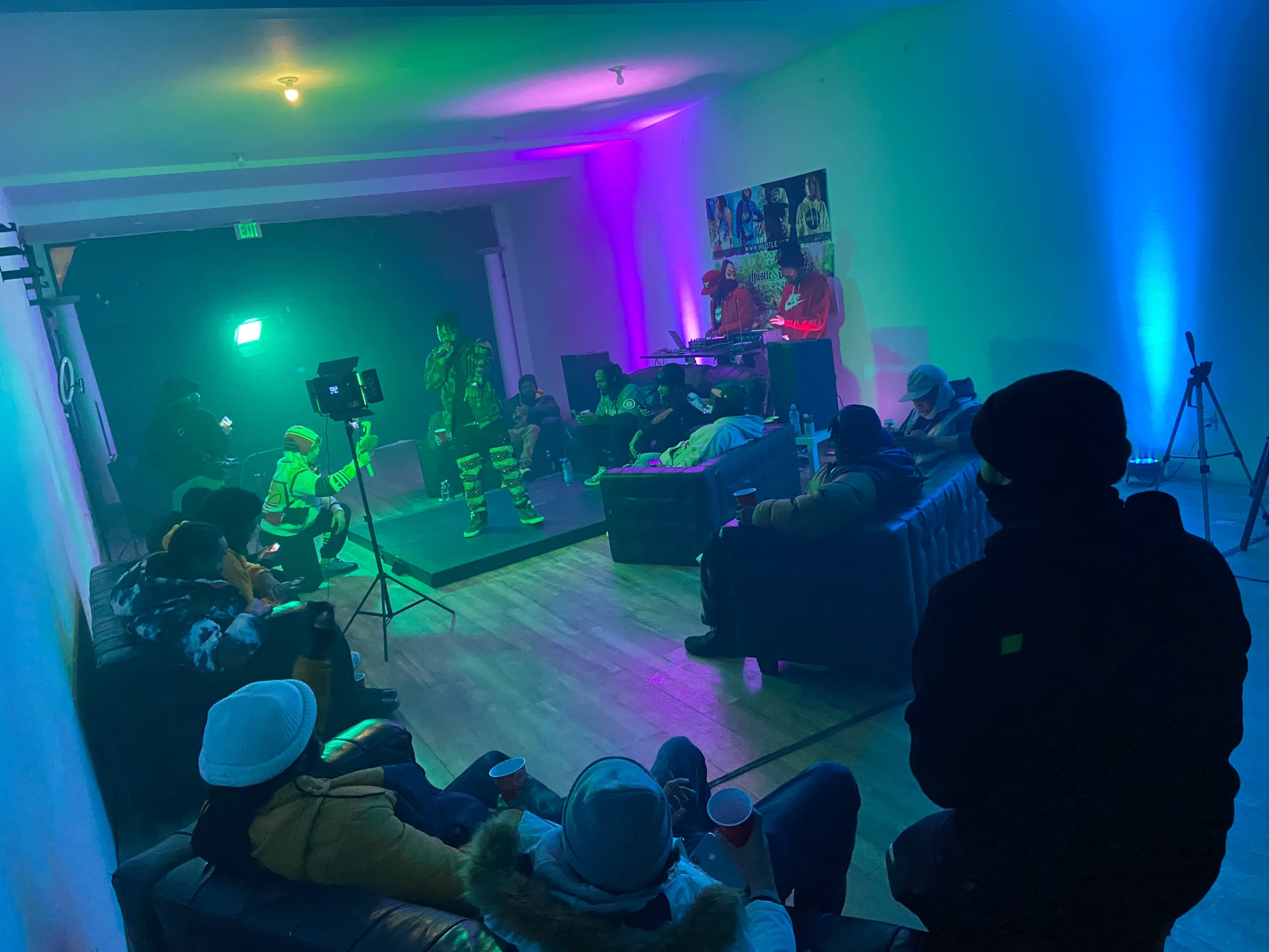 A group of people sits in a dimly lit room with colorful lights, while two performers play music on a small stage.