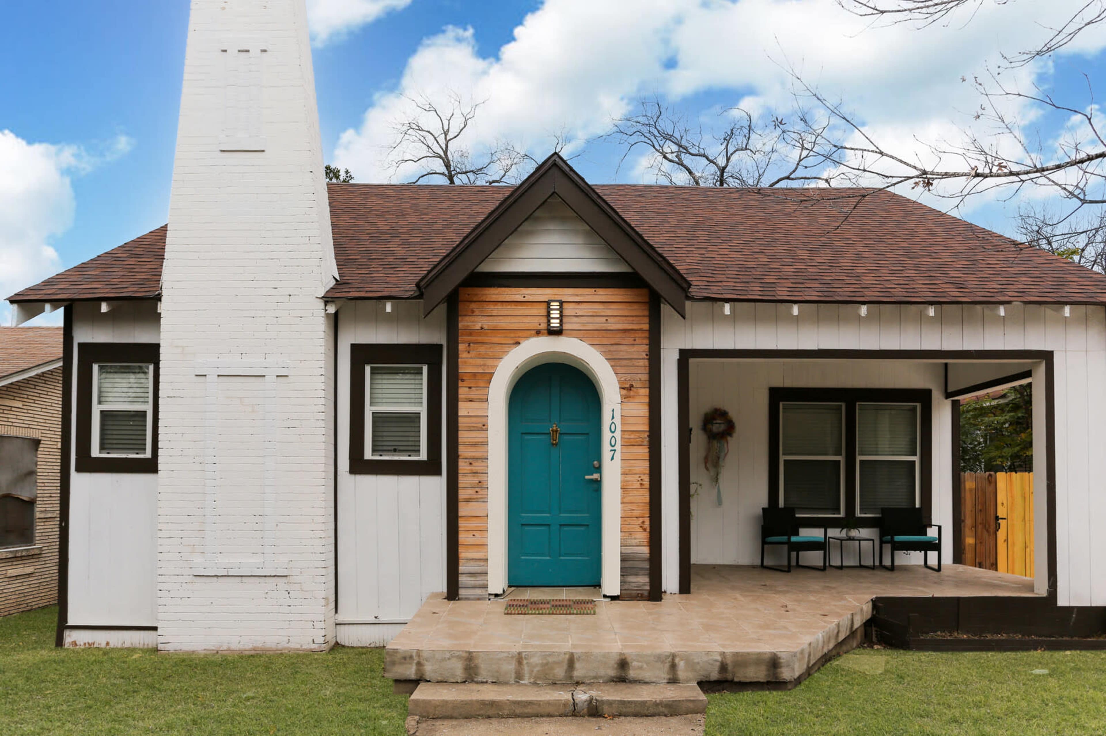 The image shows a single-story house with a white exterior, a wooden accent wall, a teal front door, and a small porch with two chairs.