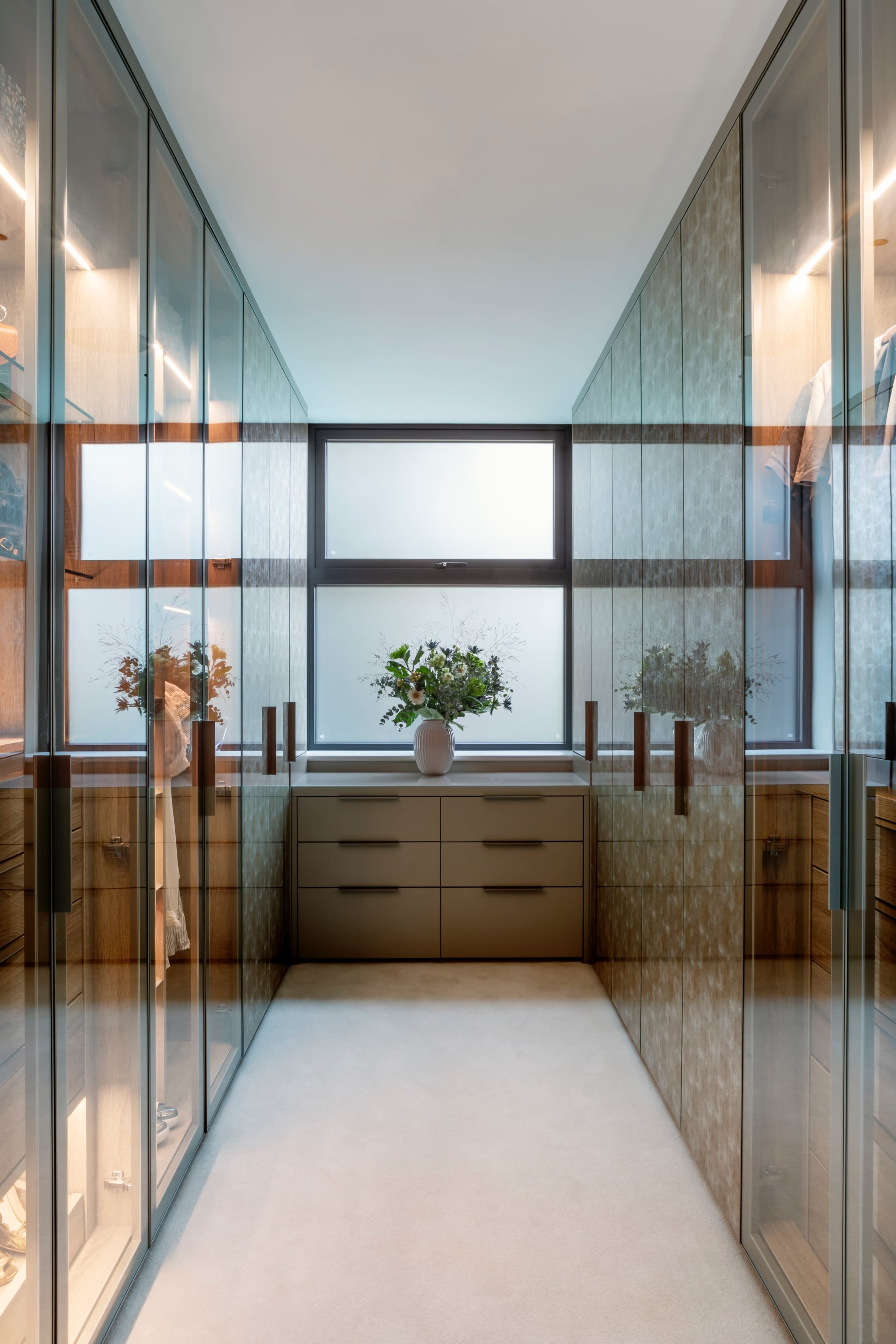 The image shows a well-organized walk-in closet with glass-fronted cabinets, neutral-colored walls, and a central display area featuring a vase of flowers.