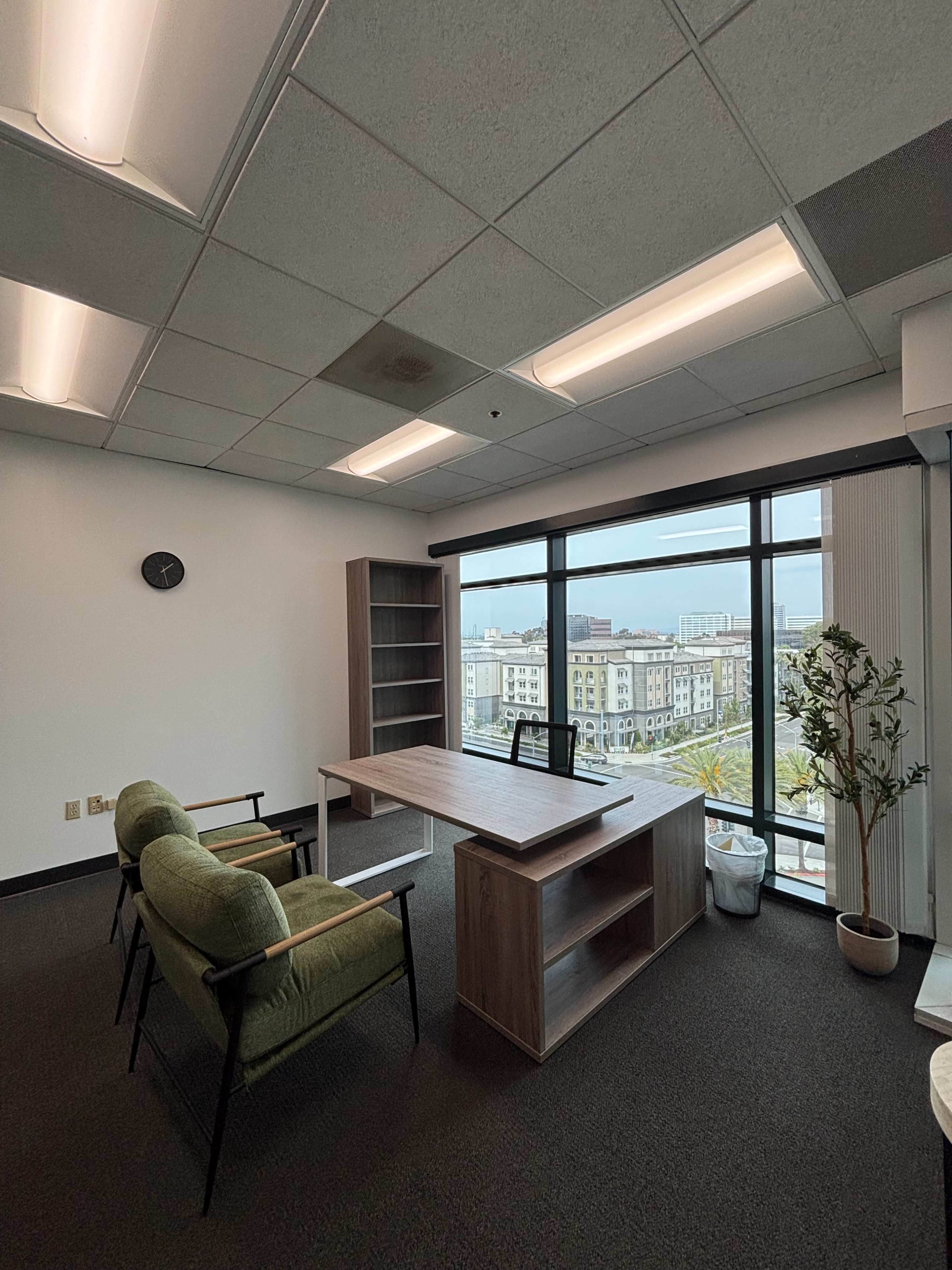 The image shows a modern office space featuring a wooden desk, two green chairs, a bookshelf, and a view of a cityscape through large windows.