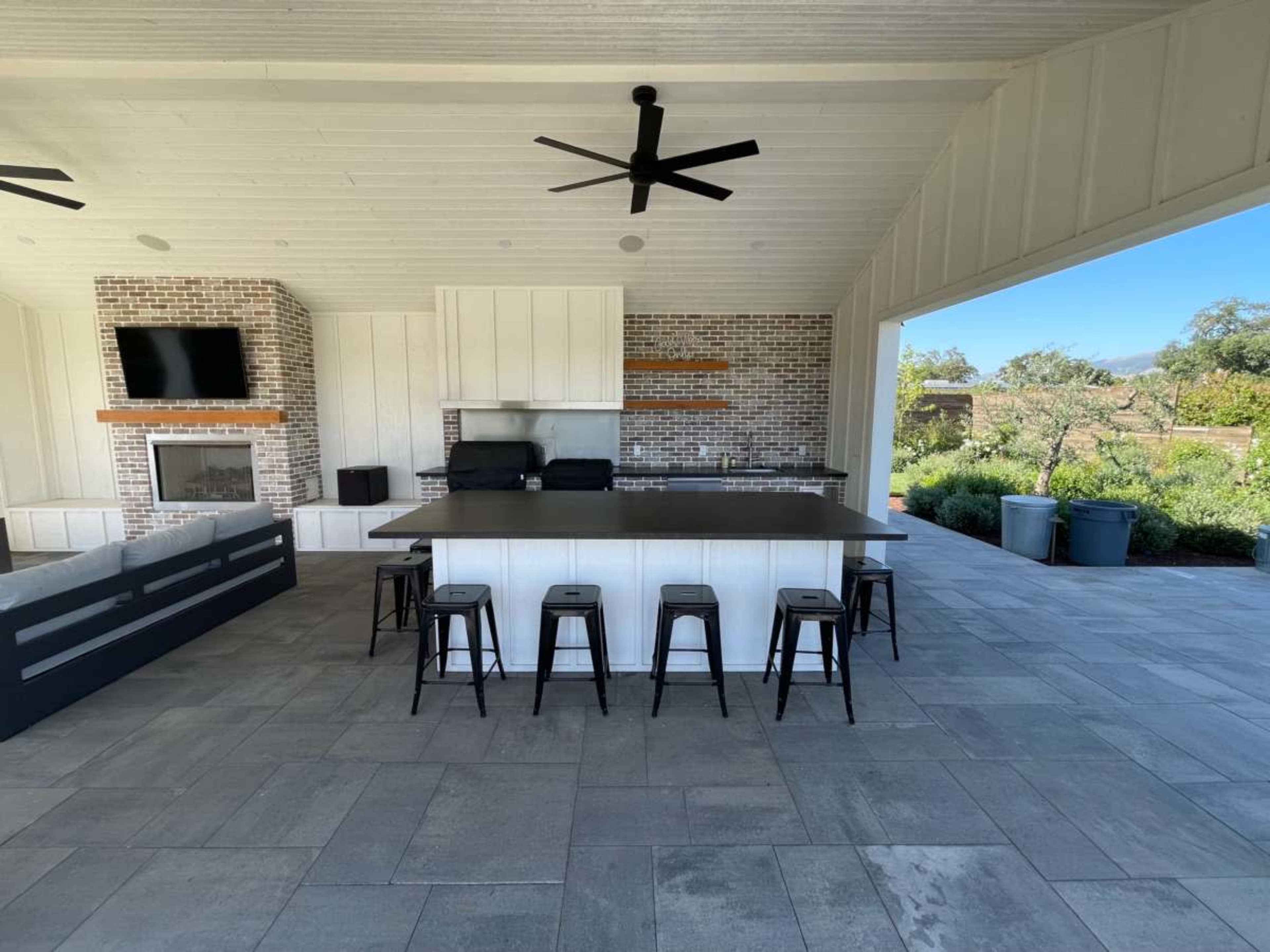 The image shows a modern outdoor kitchen with a large island, bar stools, a grill, and a stone backsplash, set in a covered patio area with an open view of the surrounding landscape.