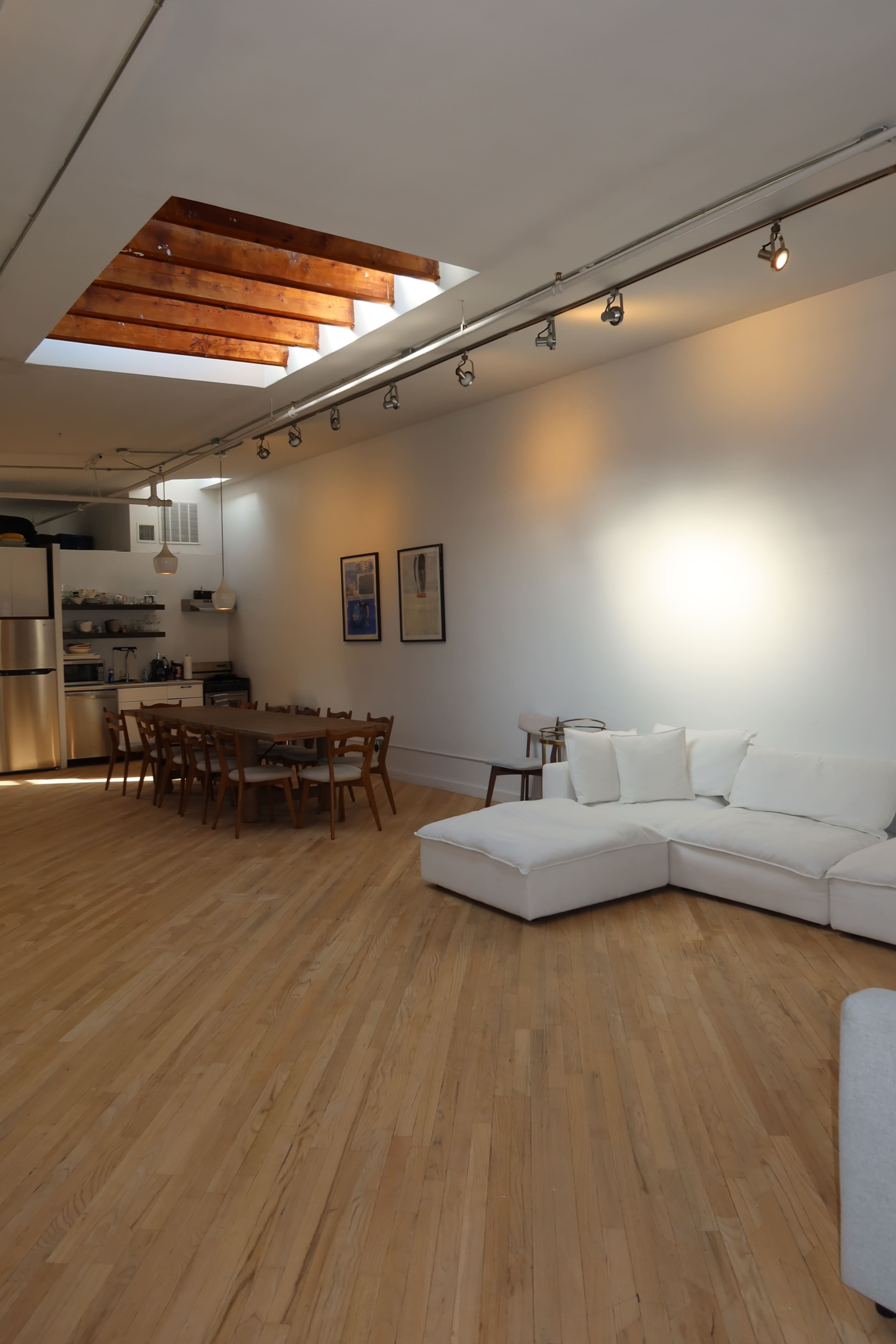The image shows a spacious, modern living area with a sectional sofa, dining table, and a skylight illuminating the space.