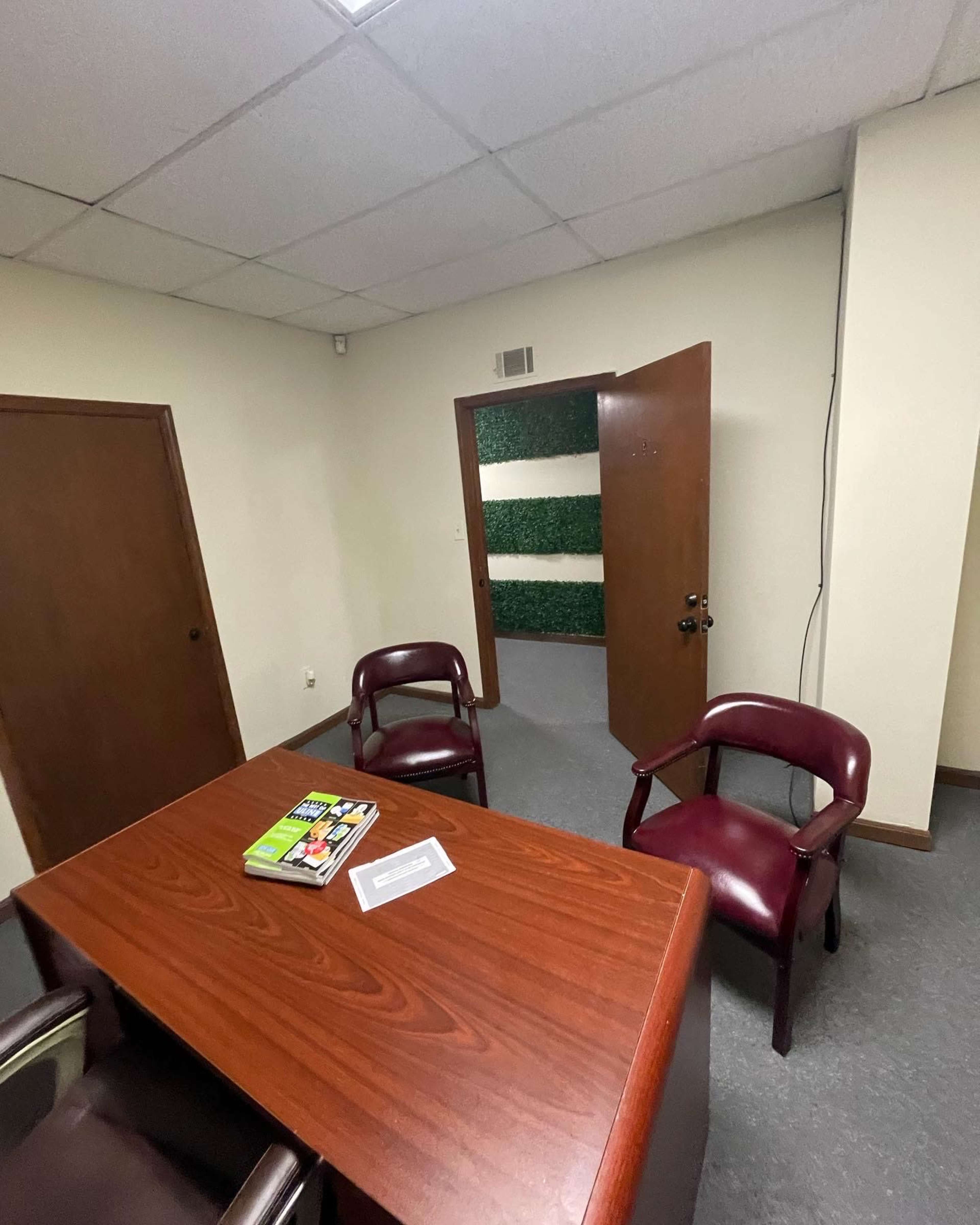 An office scene features a wooden table with a book and a paper on it, two maroon chairs, and a partially open door leading to a green wall.