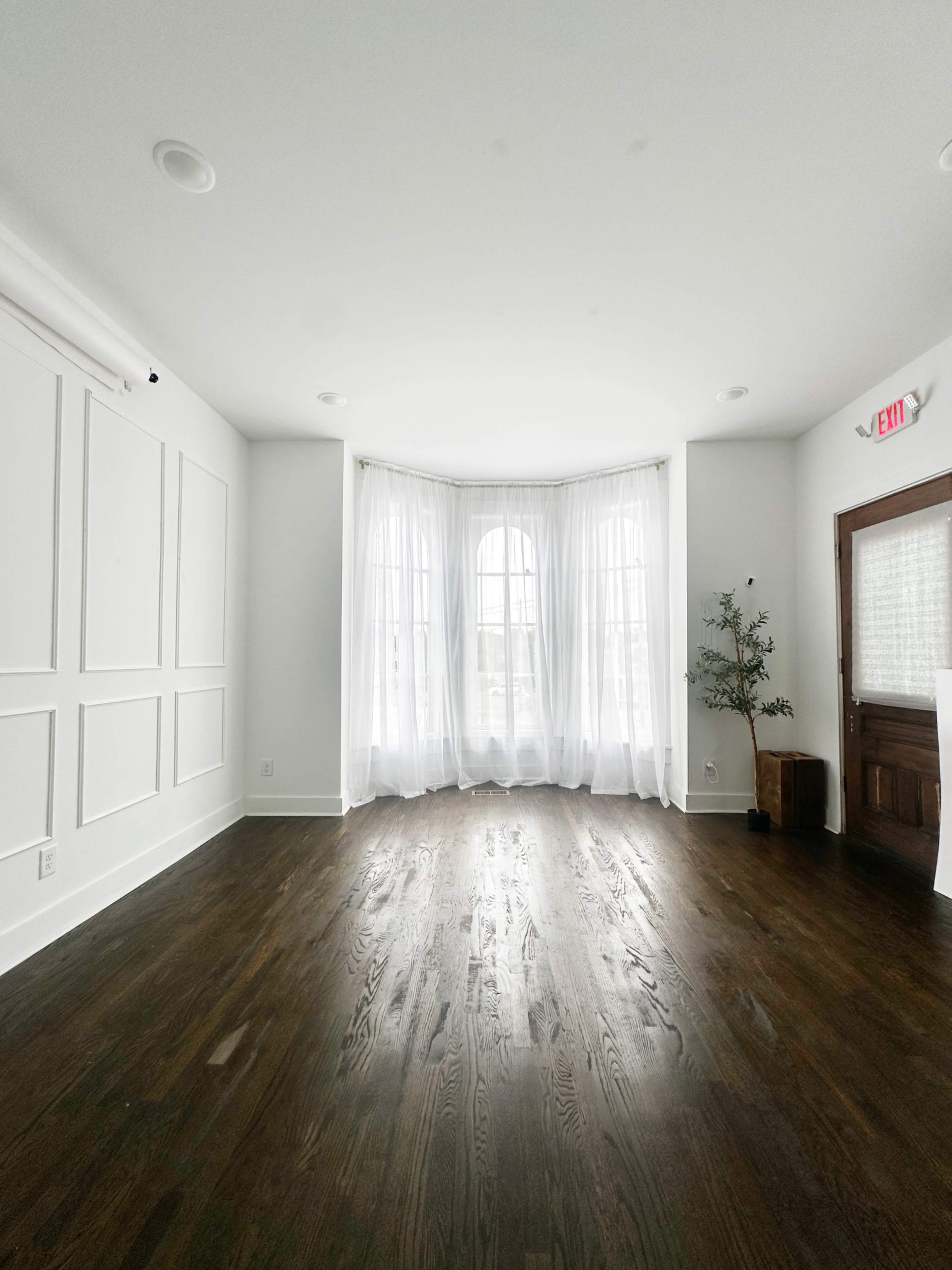 The image shows a spacious, empty room with large windows covered by sheer white curtains and a wooden floor.