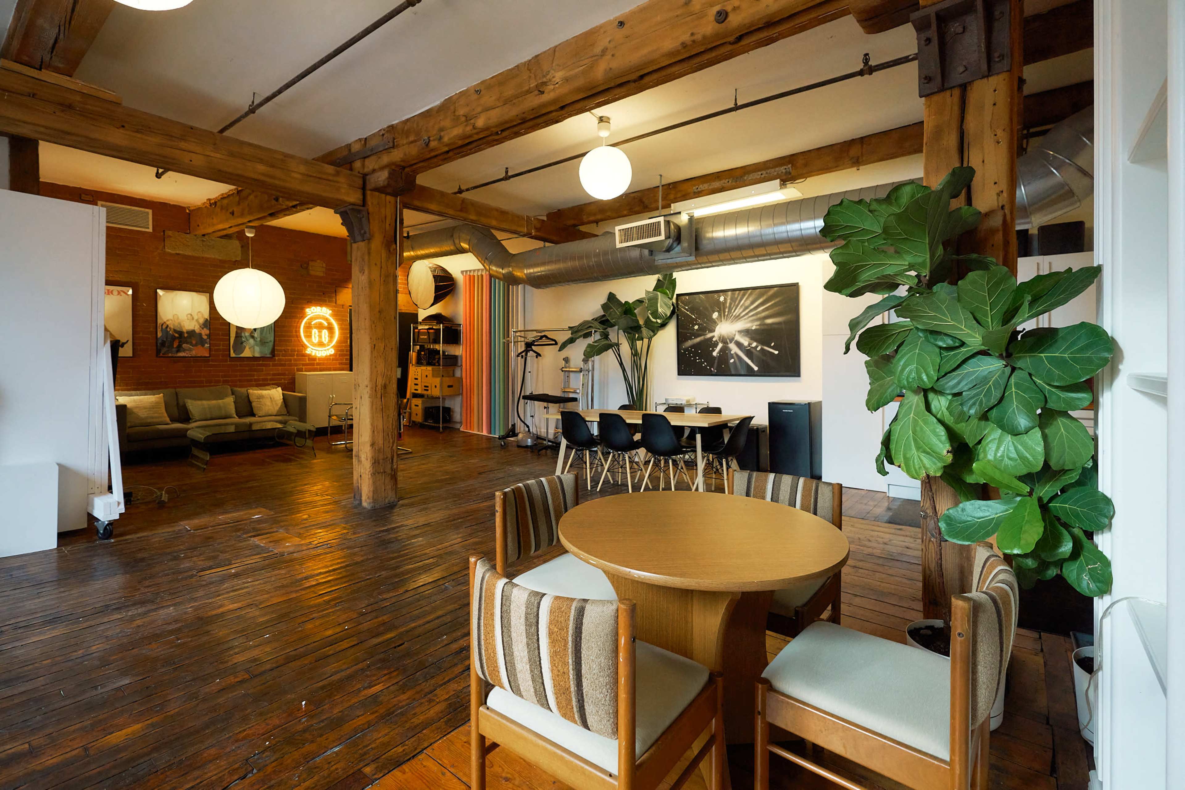 The image shows an open-style loft interior featuring wooden beams, a round dining table with chairs, a seating area with a sofa, and various plants scattered throughout.