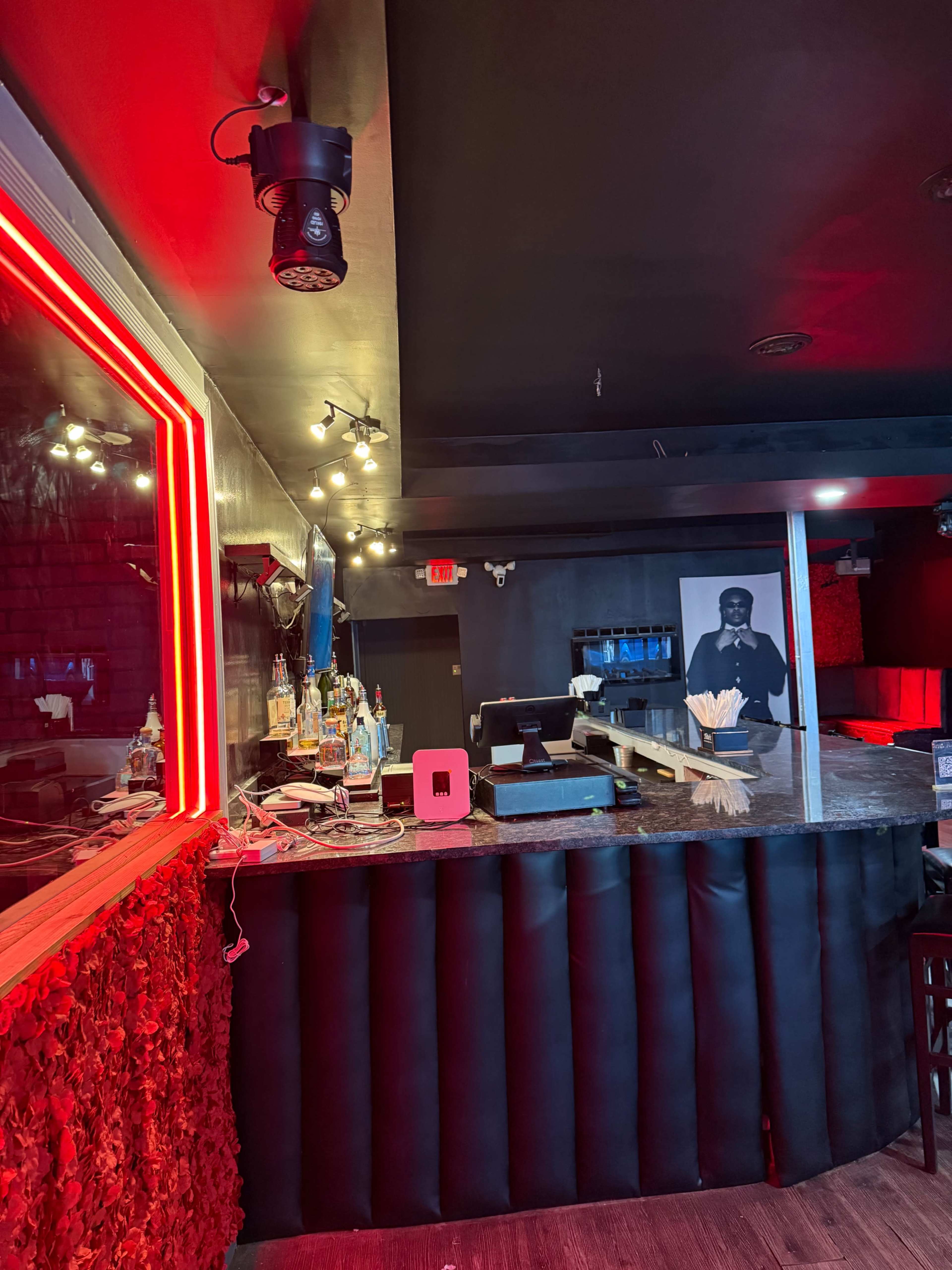 The image shows a dimly lit bar area with a granite countertop, illuminated by red lighting, and a wall decorated with a large circular mirror and a black-and-white portrait.