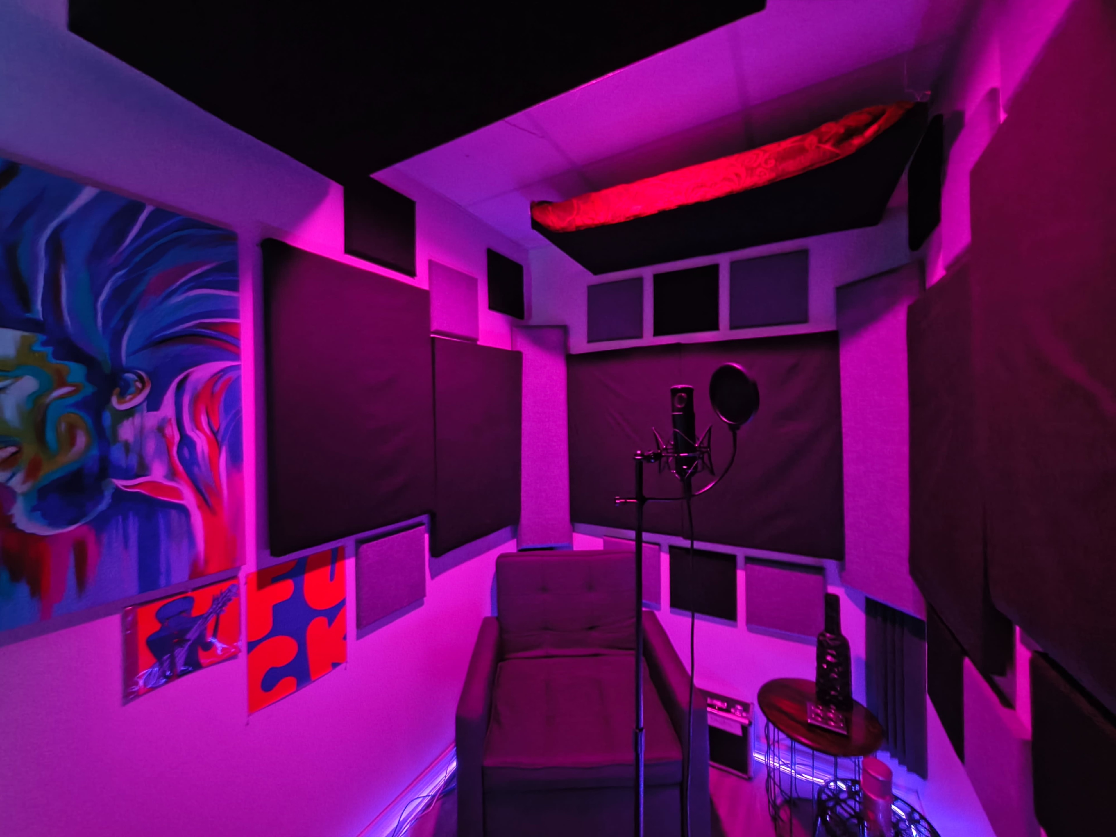 The image depicts a small music recording studio featuring soundproofing panels, a microphone on a stand, a gray couch, and purple lighting.