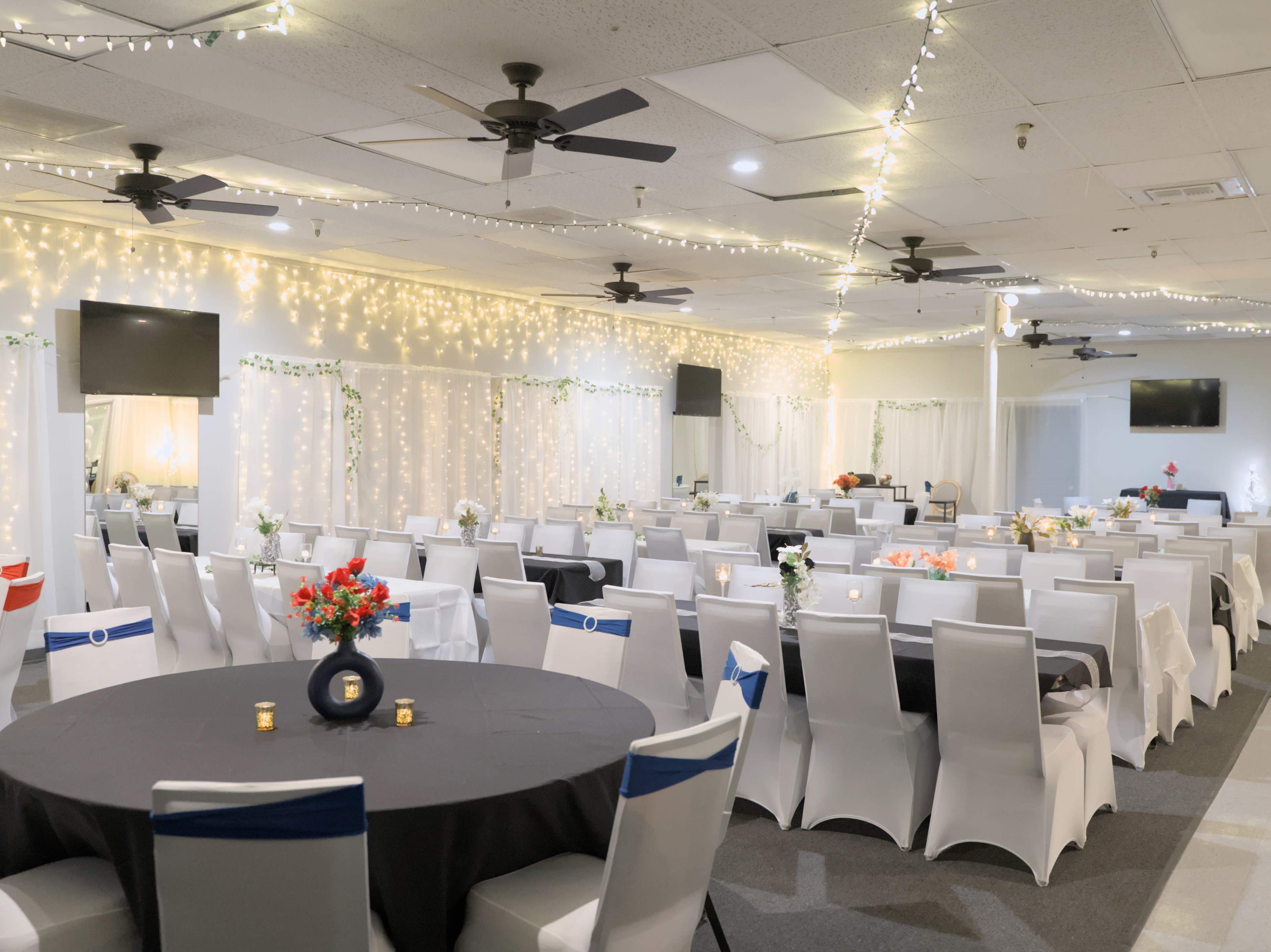 A banquet hall featuring rows of tables dressed in black and white linens, adorned with centerpieces, and illuminated by string lights overhead.