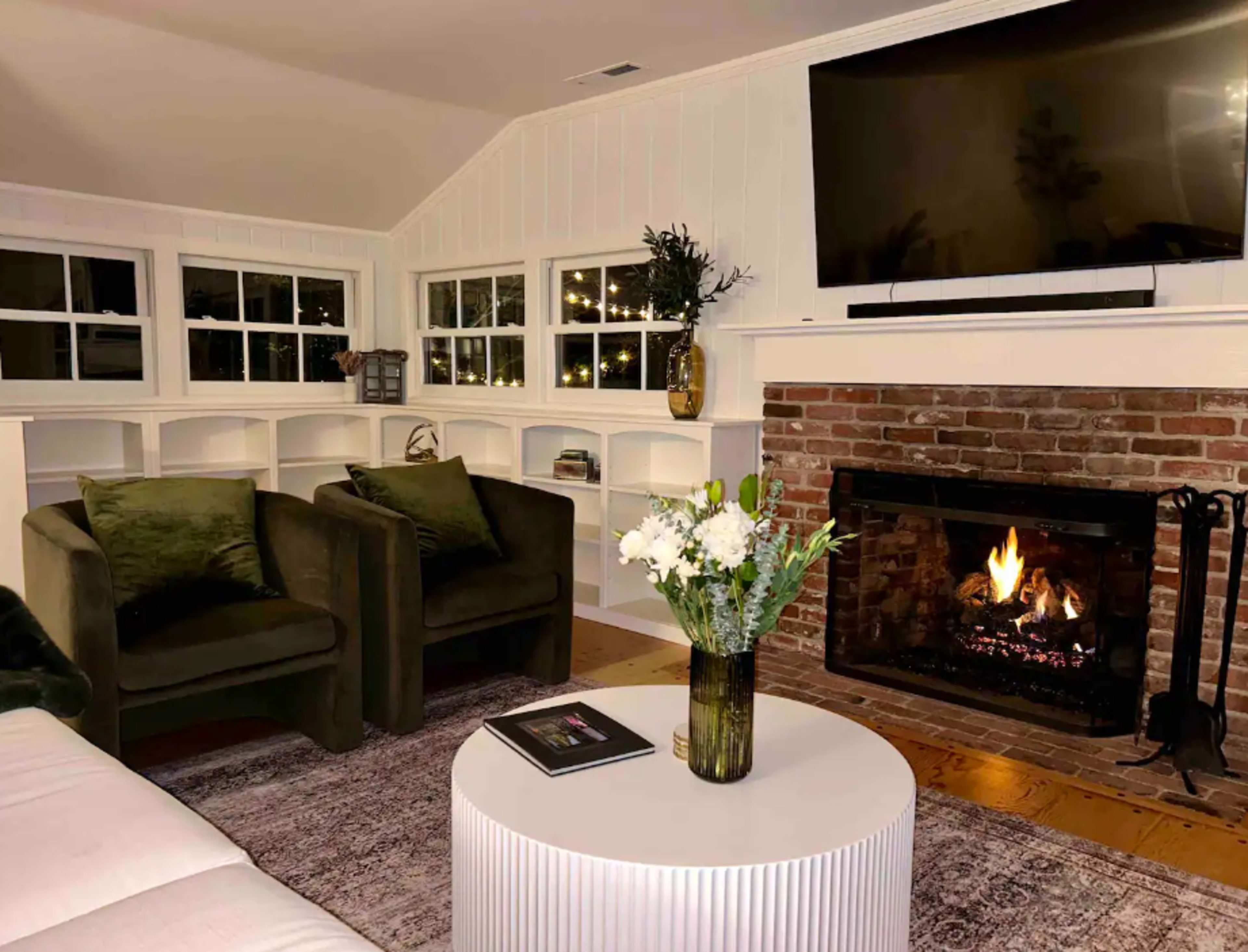 A cozy living room features two dark green chairs, a round white coffee table, and a fireplace with a fire burning, all set against a backdrop of white shiplap walls and large windows.