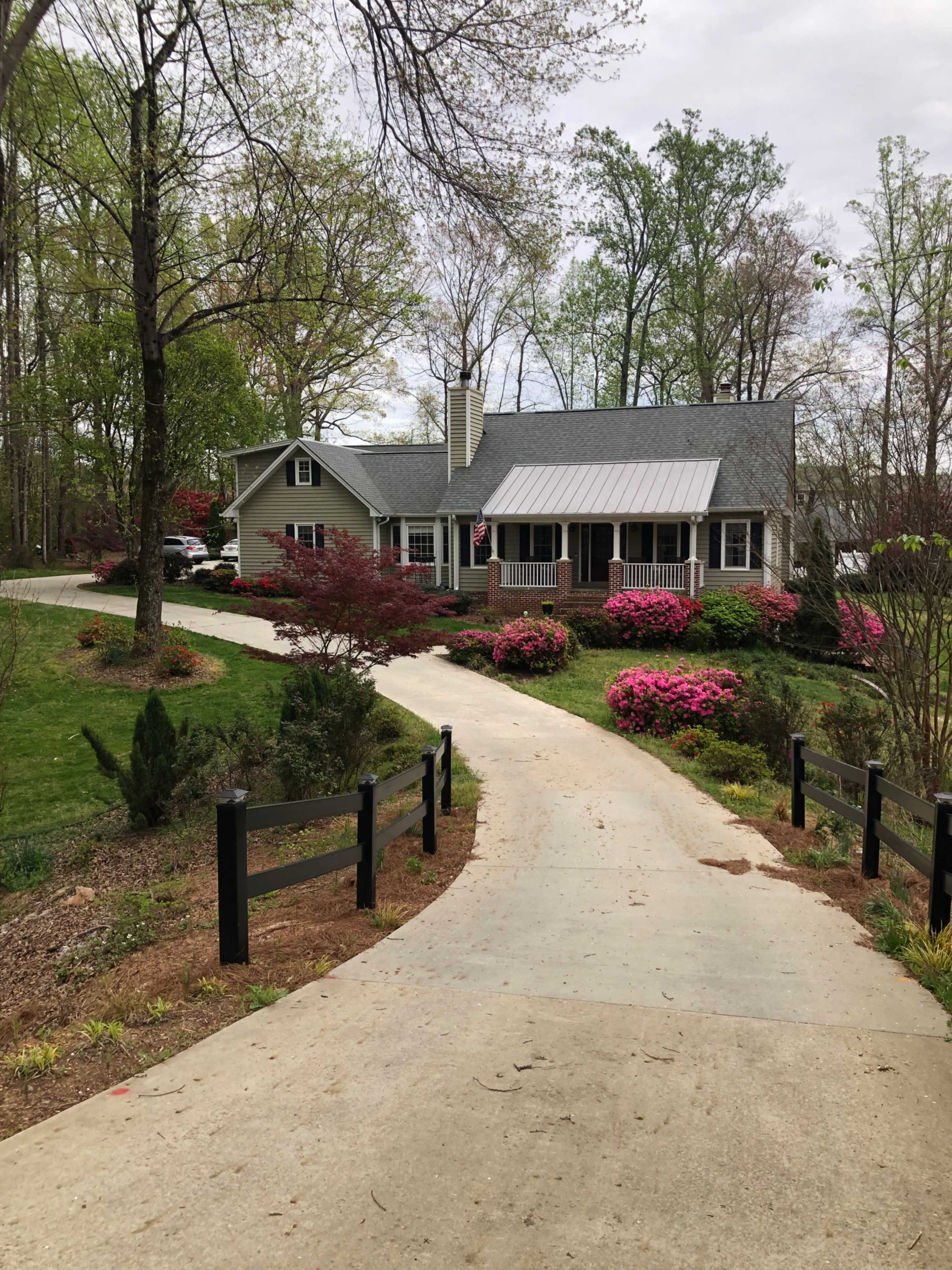 A gravel driveway leads to a single-story house surrounded by colorful flower beds and greenery.
