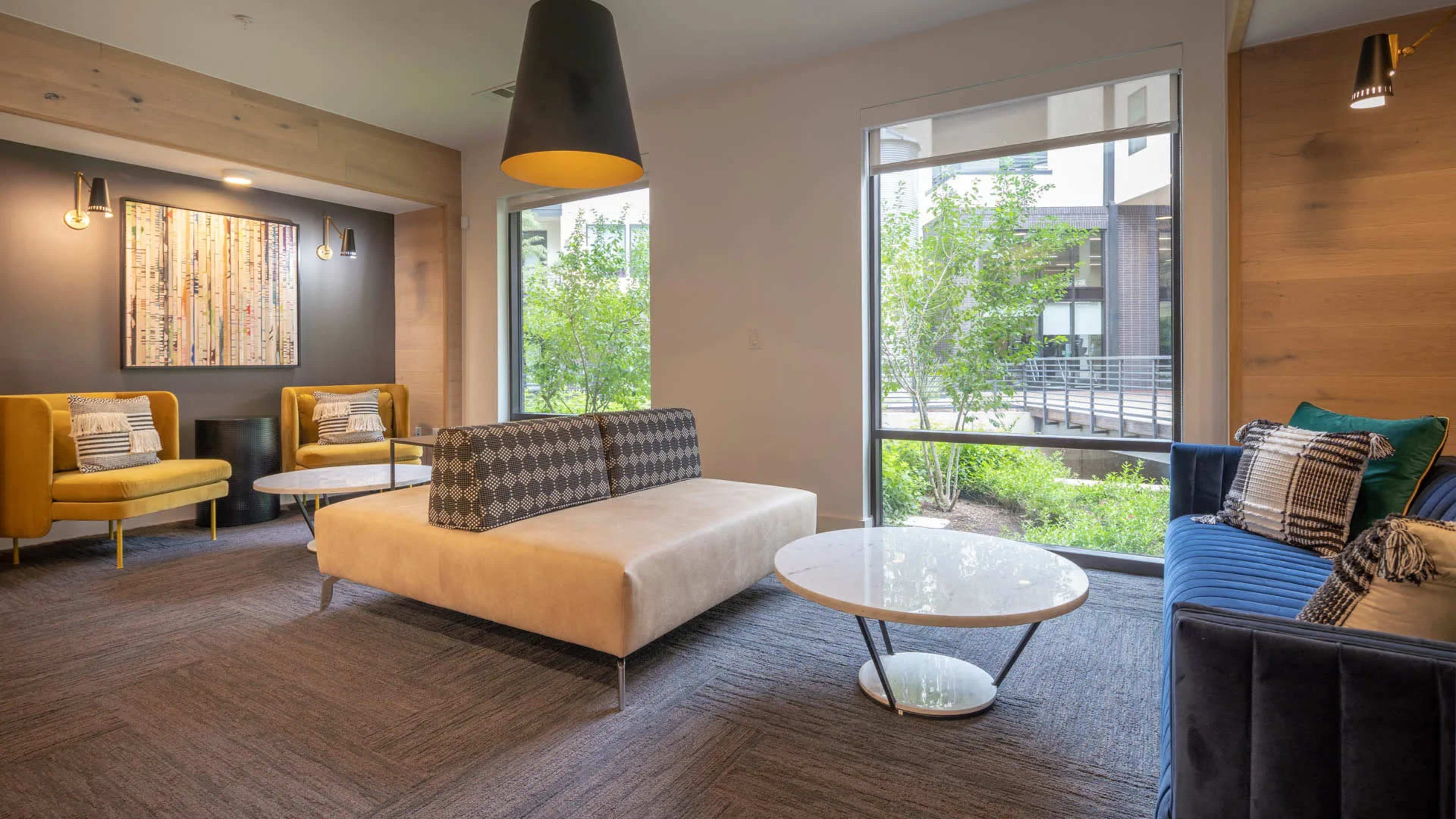 The image shows a modern lounge area with two yellow chairs, a gray sofa, and two round coffee tables near large windows that allow natural light to enter.