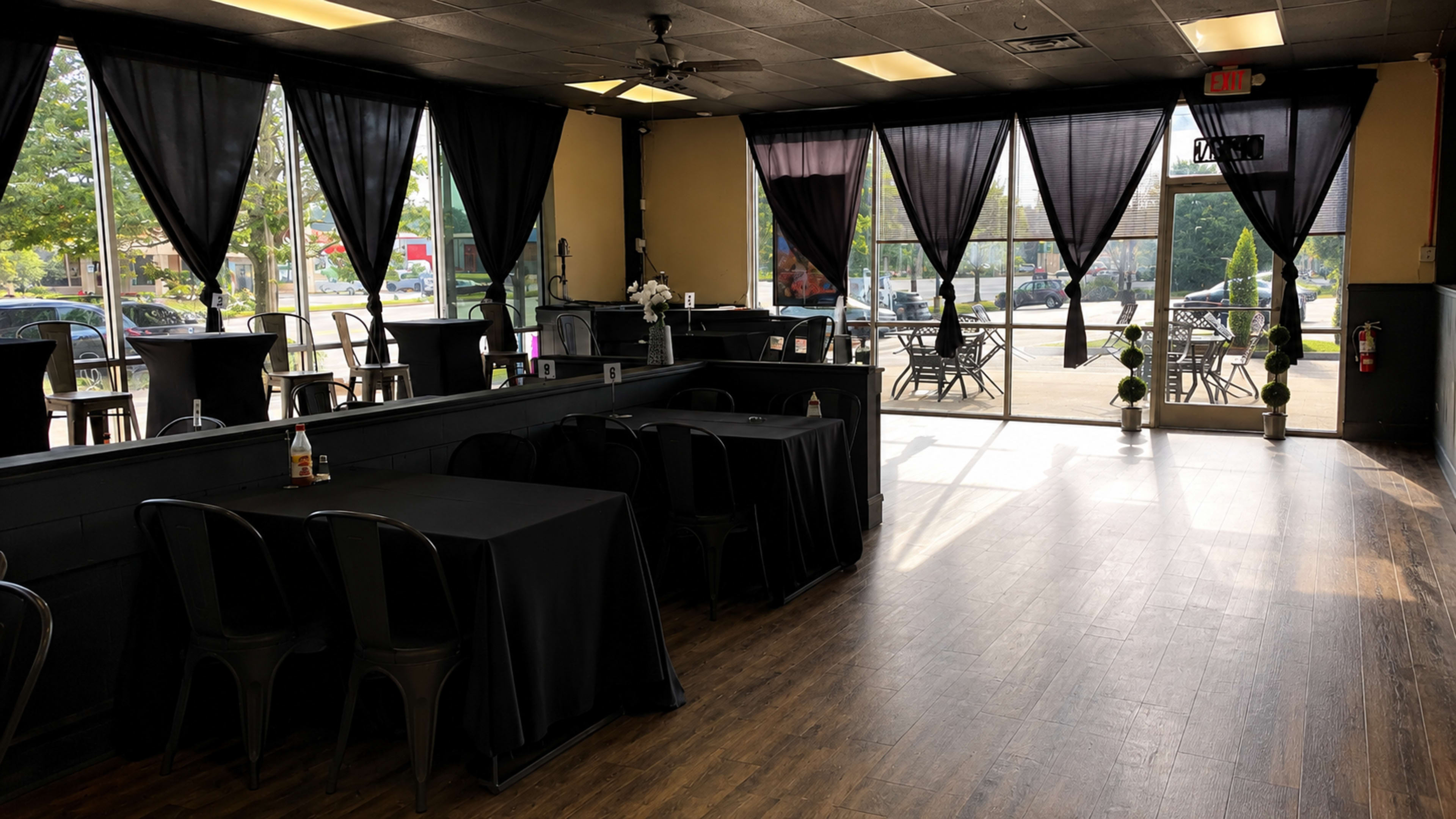 The image shows an interior of a restaurant or event space with black tablecloths on tables, large windows with black curtains, and a view of the exterior.
